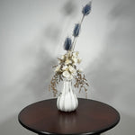 Vase of dried floral arrangement made with delicate, lace-like petals of white Cape Flowers against the intriguing, sculpted heads of blue Teasels. Adding airy lightness and enduring colour is German Statice. 