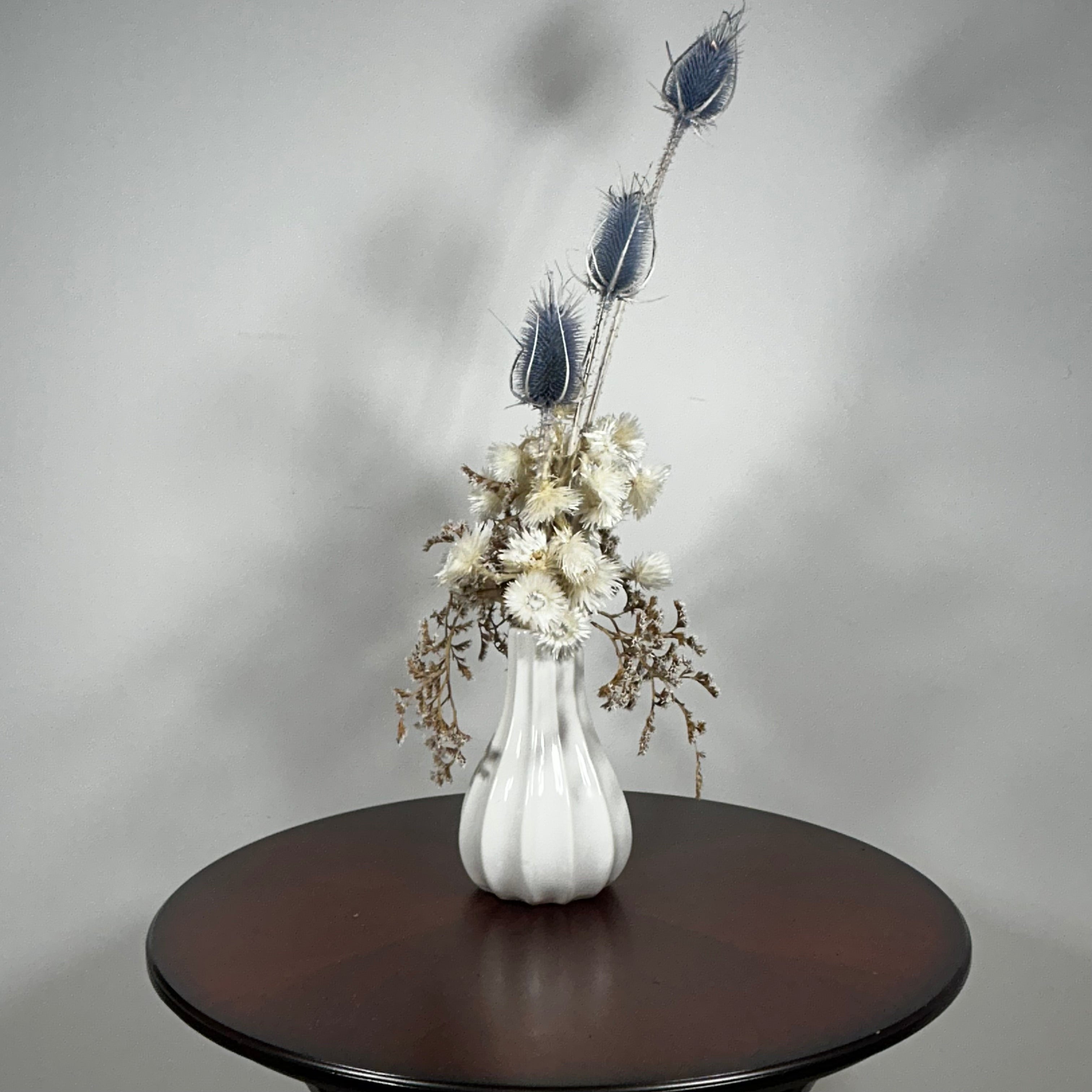 Vase of dried floral arrangement made with delicate, lace-like petals of white Cape Flowers against the intriguing, sculpted heads of blue Teasels. Adding airy lightness and enduring colour is German Statice. 
