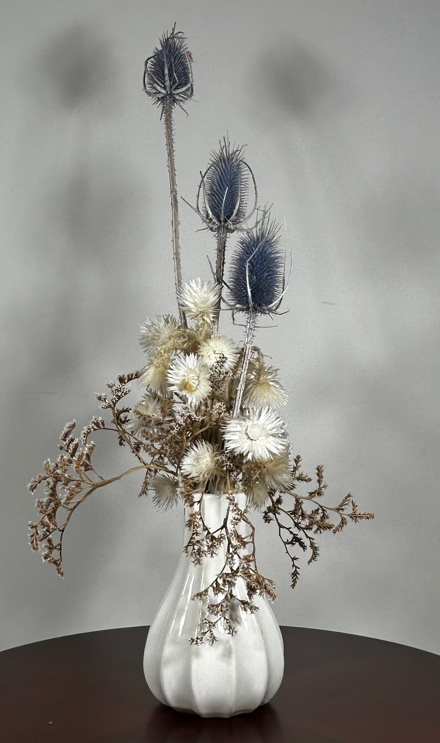 Vase of dried floral arrangement made with delicate, lace-like petals of white Cape Flowers against the intriguing, sculpted heads of blue Teasels. Adding airy lightness and enduring colour is German Statice. 