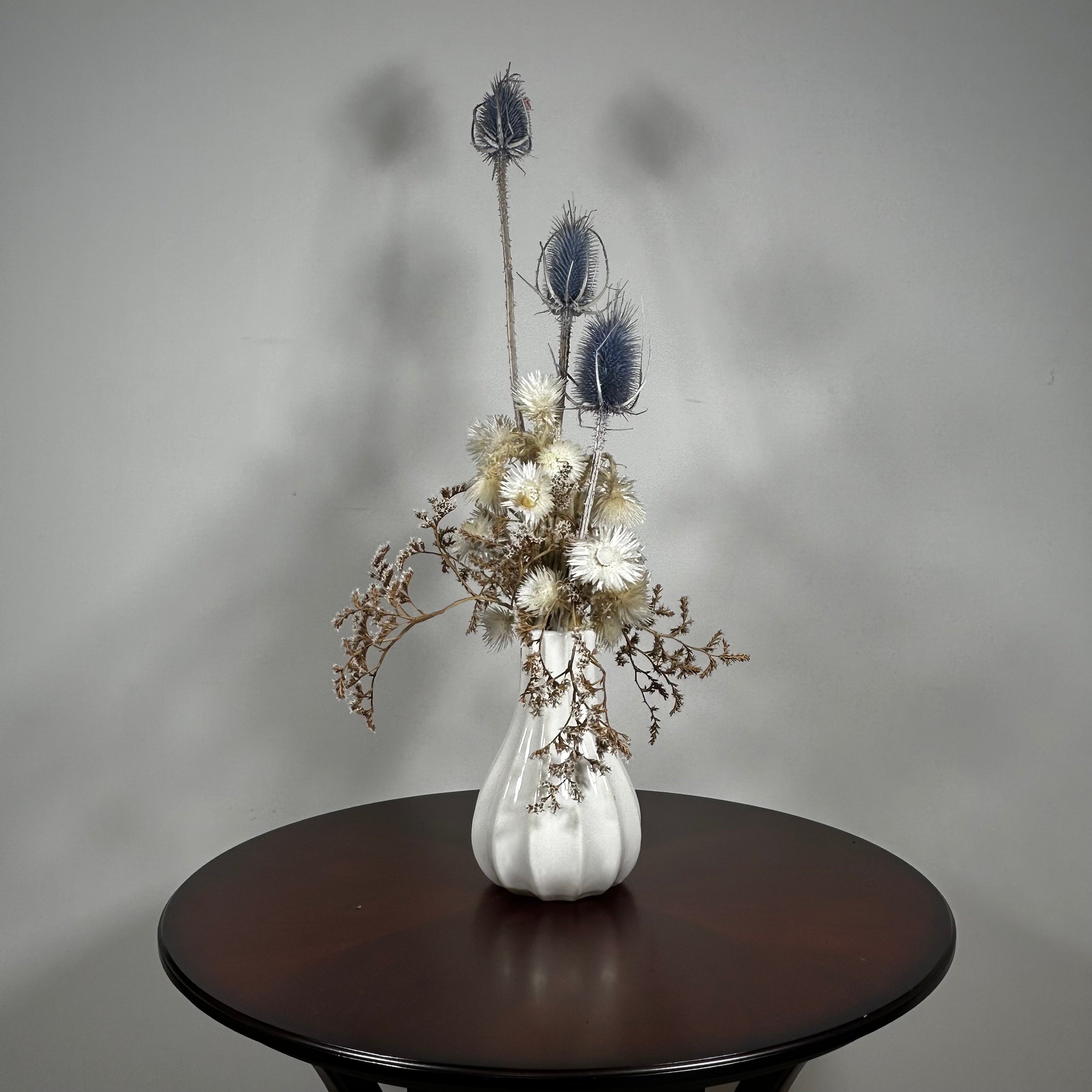Vase of dried floral arrangement made with delicate, lace-like petals of white Cape Flowers against the intriguing, sculpted heads of blue Teasels. Adding airy lightness and enduring colour is German Statice. 