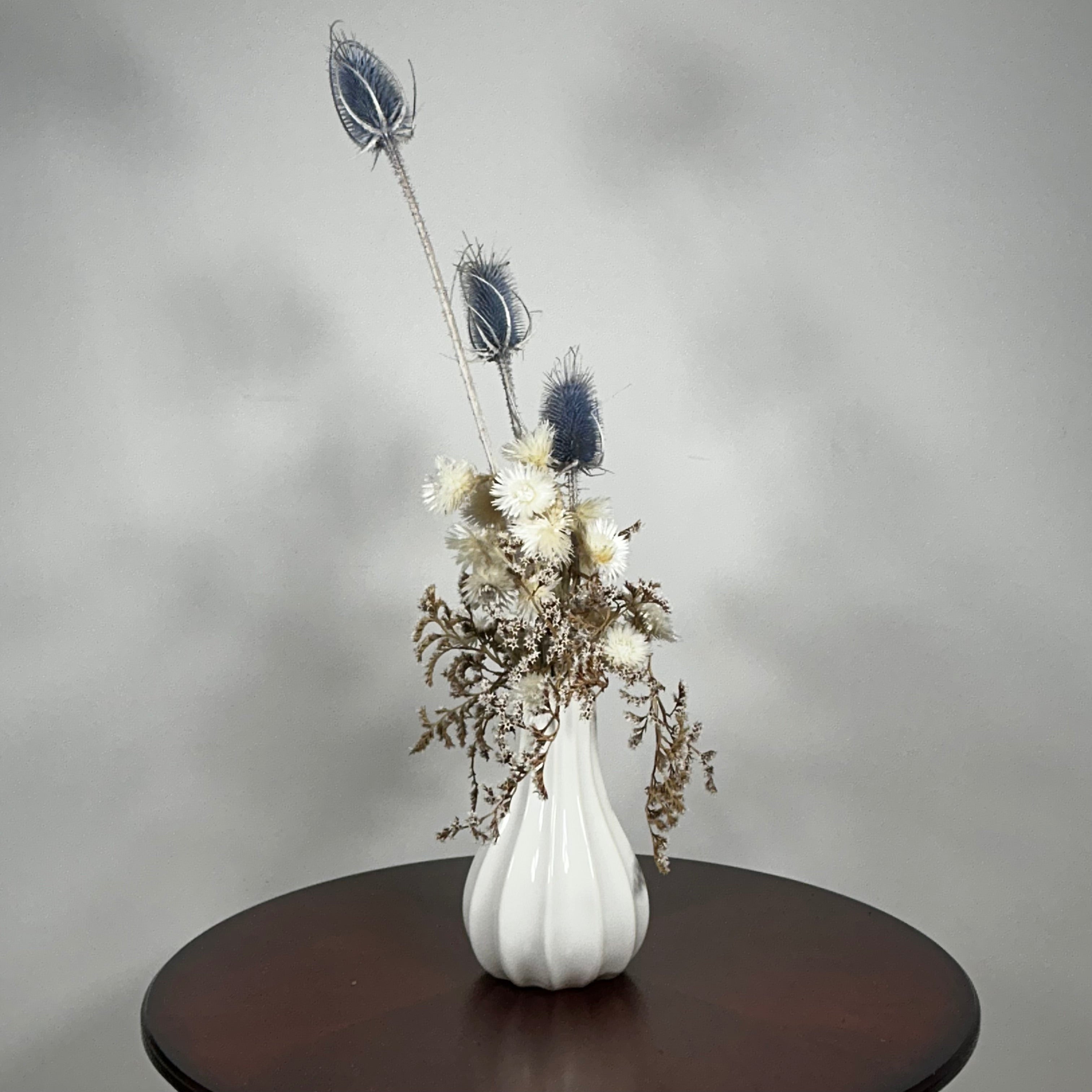 Vase of dried floral arrangement made with delicate, lace-like petals of white Cape Flowers against the intriguing, sculpted heads of blue Teasels. Adding airy lightness and enduring colour is German Statice. 