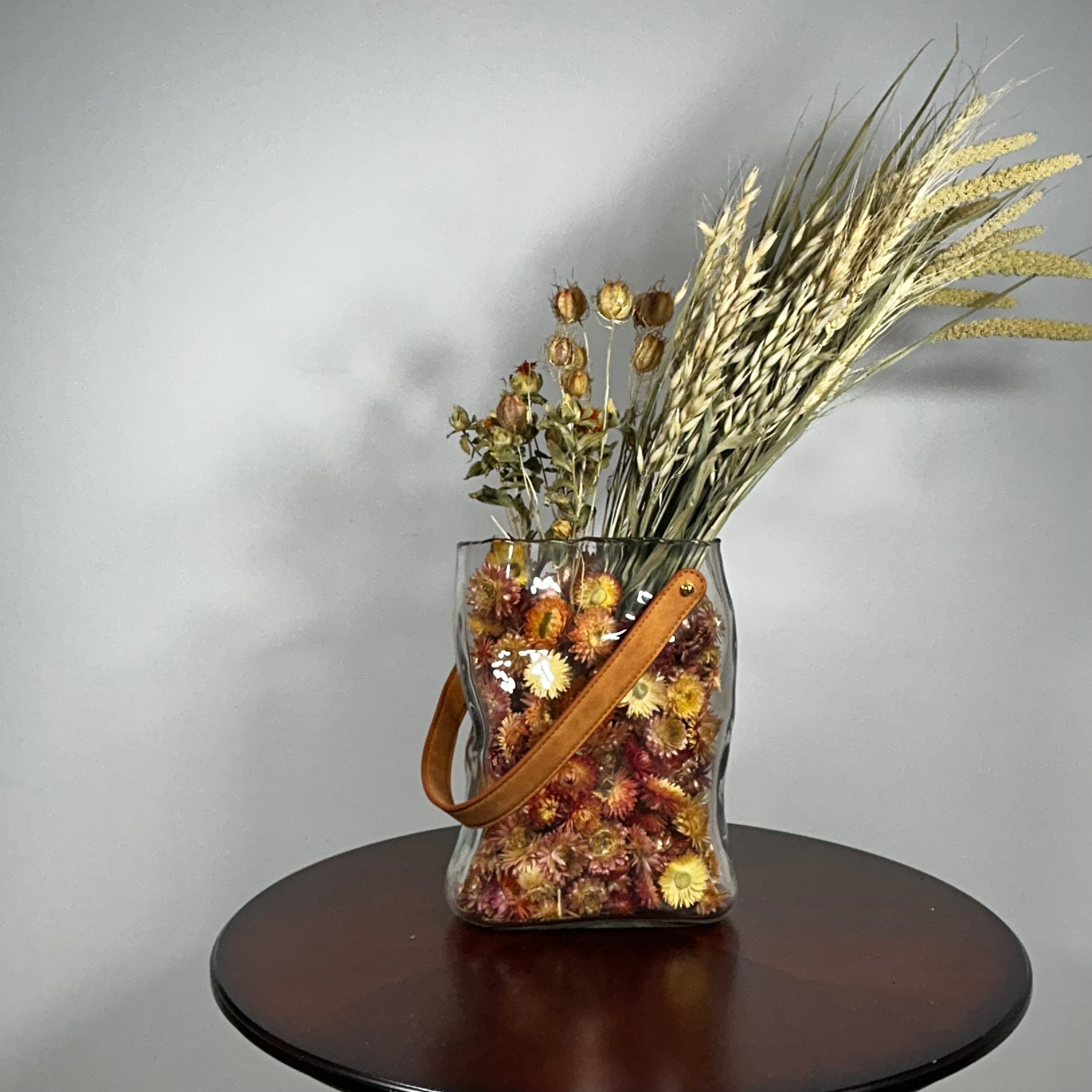 Decorative glass vase shaped like a paper bag with a leather handle filled with dried helichrysum, a beautifully arranged array of natural oats, Nigel, and safflower rise up from the vase.