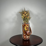 Decorative glass vase shaped like a paper bag with a leather handle filled with dried helichrysum, a beautifully arranged array of natural oats, Nigel, and safflower rise up from the vase.