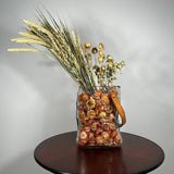 Decorative glass vase shaped like a paper bag with a leather handle filled with dried helichrysum, a beautifully arranged array of natural oats, Nigel, and safflower rise up from the vase.