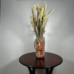Decorative glass vase shaped like a paper bag with a leather handle filled with dried helichrysum, a beautifully arranged array of natural oats, Nigel, and safflower rise up from the vase.