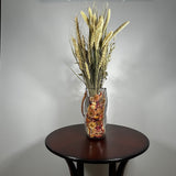 Decorative glass vase shaped like a paper bag with a leather handle filled with dried helichrysum, a beautifully arranged array of natural oats, Nigel, and safflower rise up from the vase.