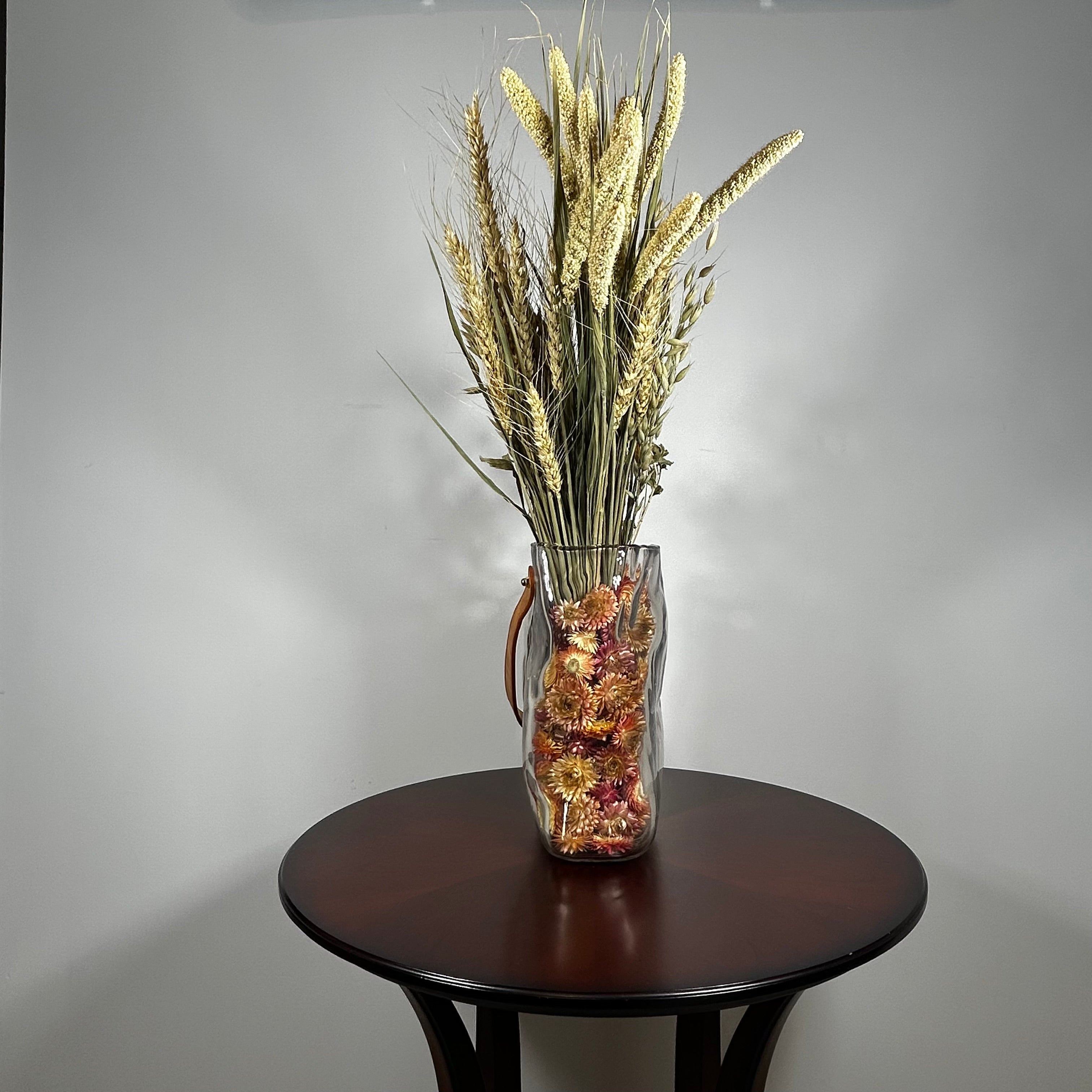 Decorative glass vase shaped like a paper bag with a leather handle filled with dried helichrysum, a beautifully arranged array of natural oats, Nigel, and safflower rise up from the vase.