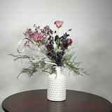 A modern white ceramic vase holds a dried flower arrangement with a delicate base of preserved asparagus ferns, from which soft plumes of gypsophila in a vibrant magenta, purple Larkspur, and soft pink peonies emerge. A final touch of playful charm is added by a single, beautifully sculpted white washed curly banana stick