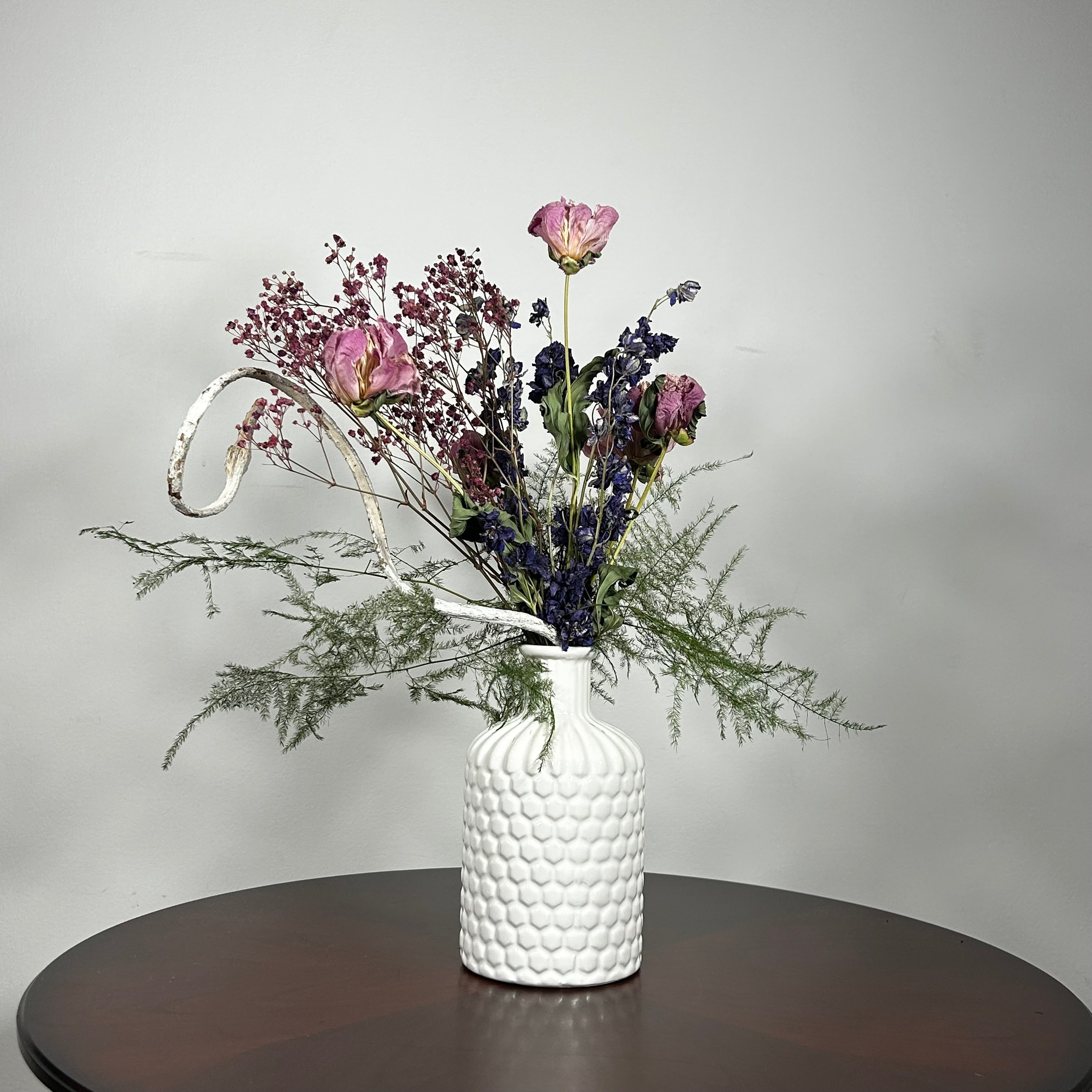 A modern white ceramic vase holds a dried flower arrangement with a delicate base of preserved asparagus ferns, from which soft plumes of gypsophila in a vibrant magenta, purple Larkspur, and soft pink peonies emerge. A final touch of playful charm is added by a single, beautifully sculpted white washed curly banana stick