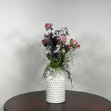 A modern white ceramic vase holds a dried flower arrangement with a delicate base of preserved asparagus ferns, from which soft plumes of gypsophila in a vibrant magenta, purple Larkspur, and soft pink peonies emerge. A final touch of playful charm is added by a single, beautifully sculpted white washed curly banana stick