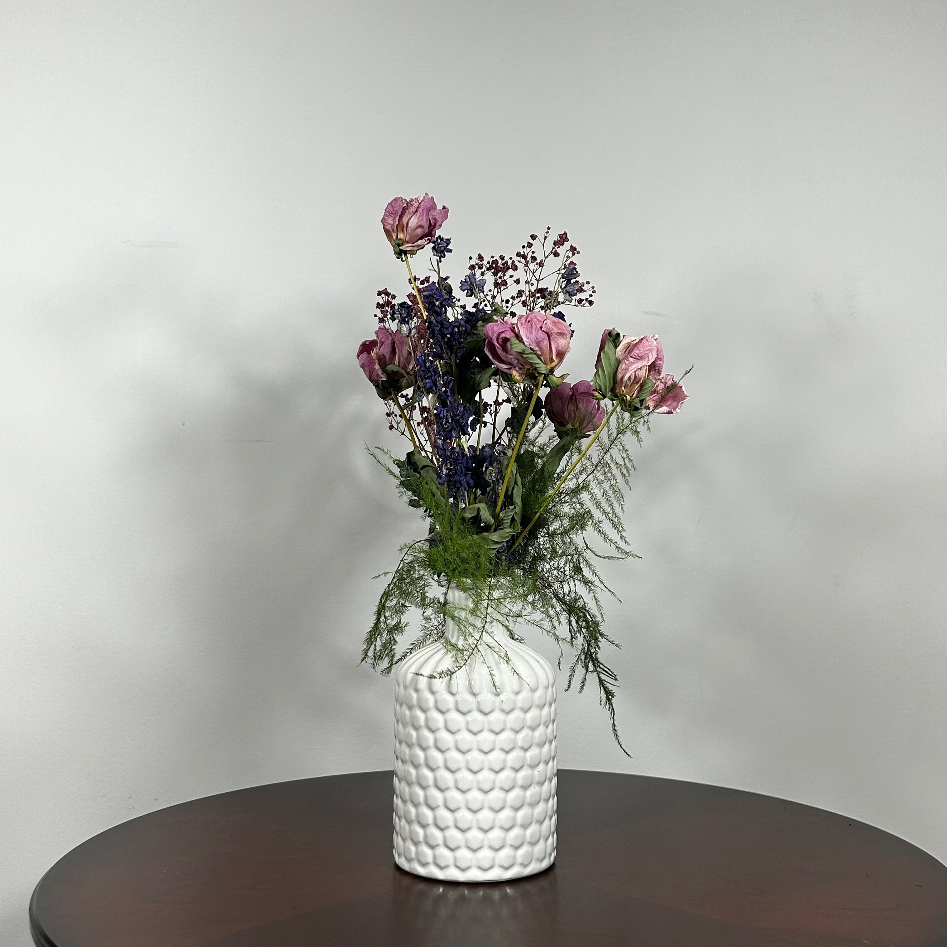 A modern white ceramic vase holds a dried flower arrangement with a delicate base of preserved asparagus ferns, from which soft plumes of gypsophila in a vibrant magenta, purple Larkspur, and soft pink peonies emerge. A final touch of playful charm is added by a single, beautifully sculpted white washed curly banana stick