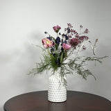 A modern white ceramic vase holds a dried flower arrangement with a delicate base of preserved asparagus ferns, from which soft plumes of gypsophila in a vibrant magenta, purple Larkspur, and soft pink peonies emerge. A final touch of playful charm is added by a single, beautifully sculpted white washed curly banana stick