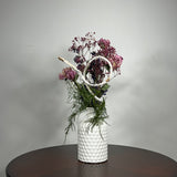 A modern white ceramic vase holds a dried flower arrangement with a delicate base of preserved asparagus ferns, from which soft plumes of gypsophila in a vibrant magenta, purple Larkspur, and soft pink peonies emerge. A final touch of playful charm is added by a single, beautifully sculpted white washed curly banana stick