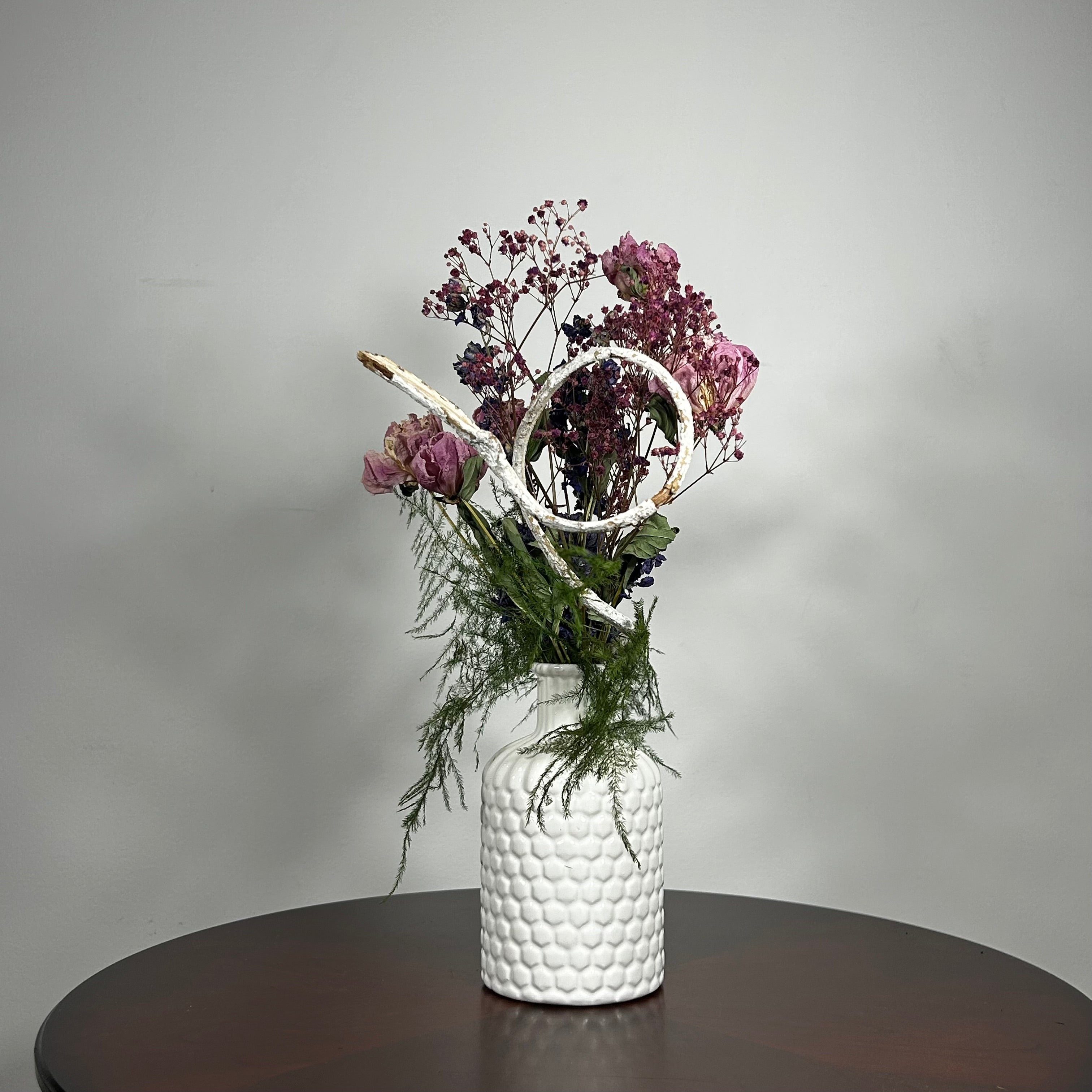 A modern white ceramic vase holds a dried flower arrangement with a delicate base of preserved asparagus ferns, from which soft plumes of gypsophila in a vibrant magenta, purple Larkspur, and soft pink peonies emerge. A final touch of playful charm is added by a single, beautifully sculpted white washed curly banana stick