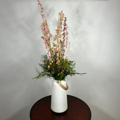 A white ceramic vase with a leather handle acts a the vessel from which a beautiful wisp of blooms rises. The central feature is the delicate spires of natural pink Larkspur, reaching upwards with gentle movement. These are complemented by charming clusters of natural pink roses and a base of lush, preserved asparagus greens. Cheerful accents of sunny yellow Statice complete this delightful composition, capturing the essence of a blossoming garden.