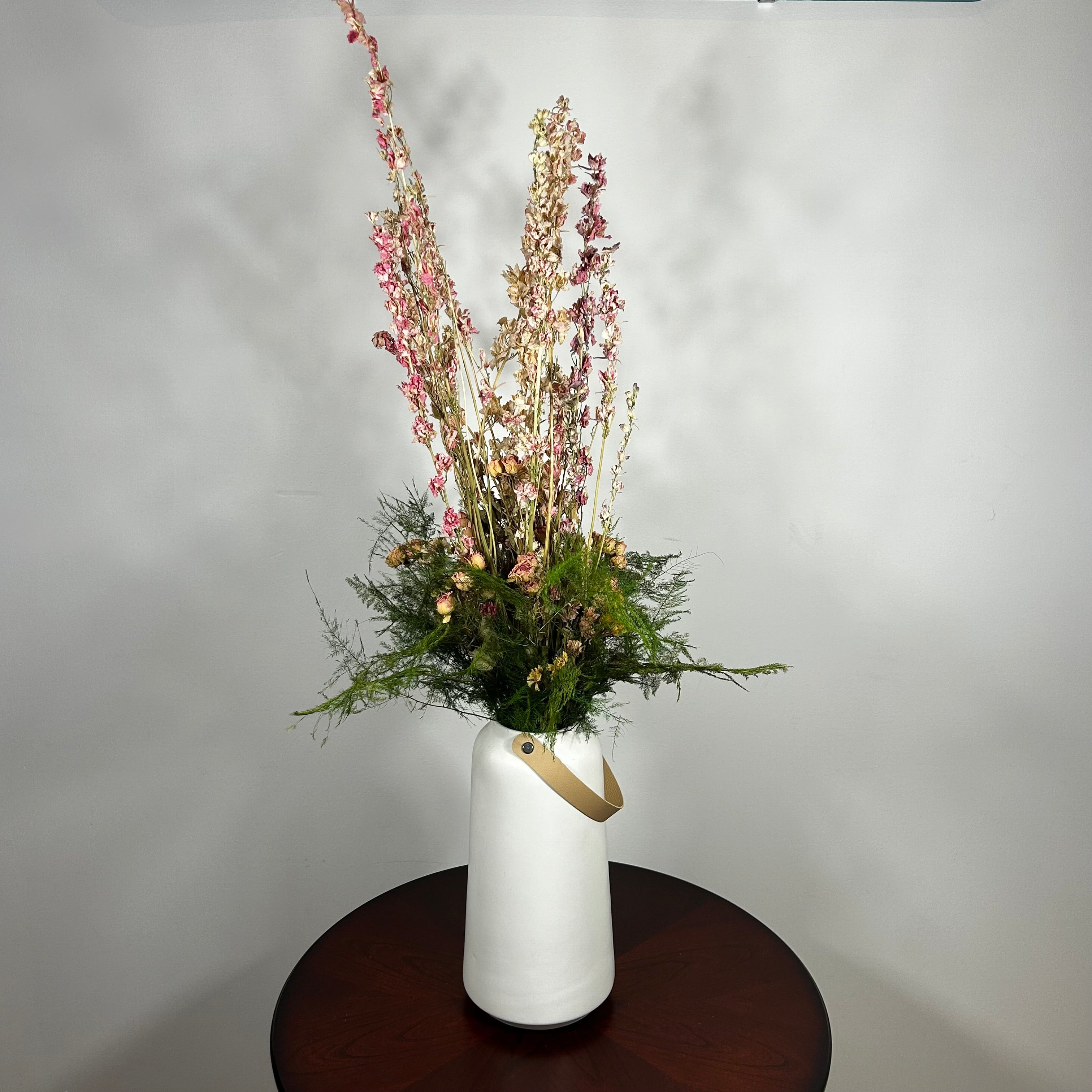 A white ceramic vase with a leather handle acts a the vessel from which a beautiful wisp of blooms rises. The central feature is the delicate spires of natural pink Larkspur, reaching upwards with gentle movement. These are complemented by charming clusters of natural pink roses and a base of lush, preserved asparagus greens. Cheerful accents of sunny yellow Statice complete this delightful composition, capturing the essence of a blossoming garden.