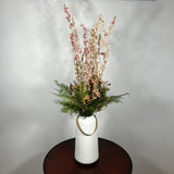 A white ceramic vase with a leather handle acts a the vessel from which a beautiful wisp of blooms rises. The central feature is the delicate spires of natural pink Larkspur, reaching upwards with gentle movement. These are complemented by charming clusters of natural pink roses and a base of lush, preserved asparagus greens. Cheerful accents of sunny yellow Statice complete this delightful composition, capturing the essence of a blossoming garden.