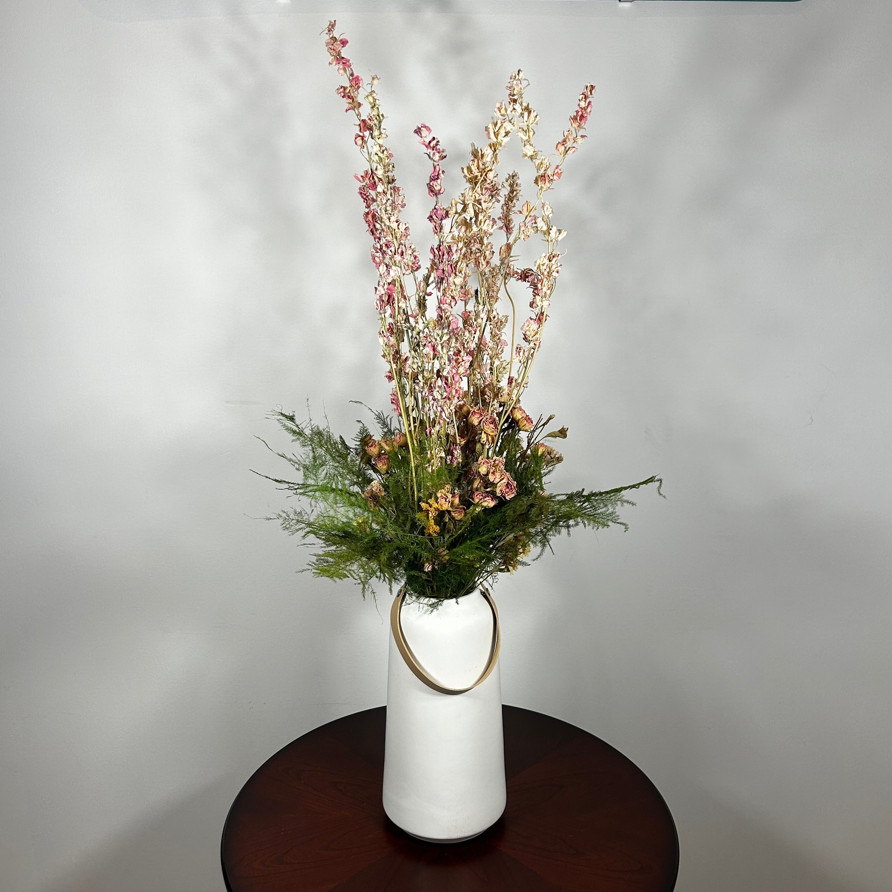 A white ceramic vase with a leather handle acts a the vessel from which a beautiful wisp of blooms rises. The central feature is the delicate spires of natural pink Larkspur, reaching upwards with gentle movement. These are complemented by charming clusters of natural pink roses and a base of lush, preserved asparagus greens. Cheerful accents of sunny yellow Statice complete this delightful composition, capturing the essence of a blossoming garden.