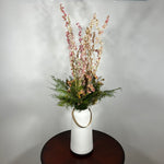 A white ceramic vase with a leather handle acts a the vessel from which a beautiful wisp of blooms rises. The central feature is the delicate spires of natural pink Larkspur, reaching upwards with gentle movement. These are complemented by charming clusters of natural pink roses and a base of lush, preserved asparagus greens. Cheerful accents of sunny yellow Statice complete this delightful composition, capturing the essence of a blossoming garden. 3