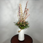 A white ceramic vase with a leather handle acts a the vessel from which a beautiful wisp of blooms rises. The central feature is the delicate spires of natural pink Larkspur, reaching upwards with gentle movement. These are complemented by charming clusters of natural pink roses and a base of lush, preserved asparagus greens. Cheerful accents of sunny yellow Statice complete this delightful composition, capturing the essence of a blossoming garden. 4