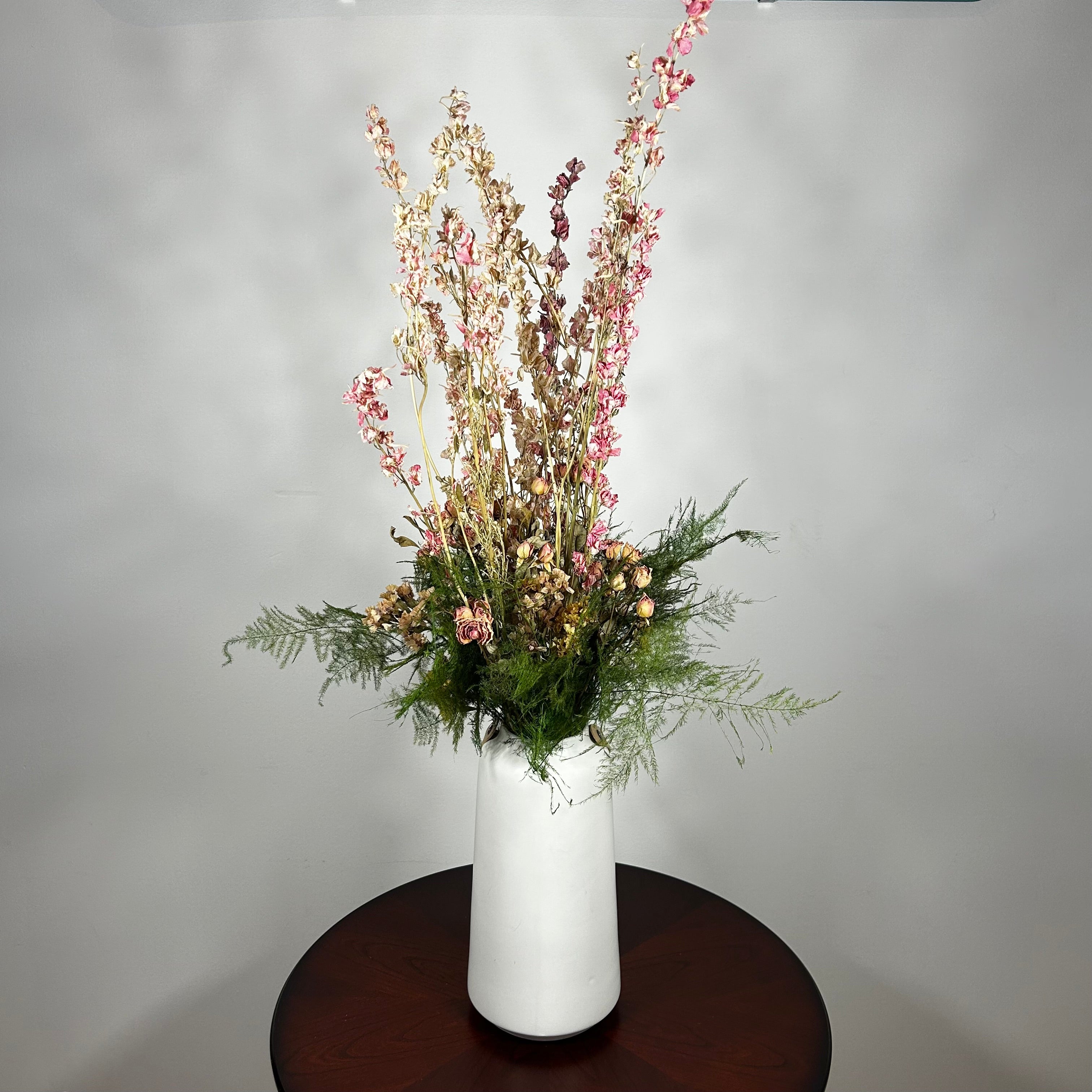 A white ceramic vase with a leather handle acts a the vessel from which a beautiful wisp of blooms rises. The central feature is the delicate spires of natural pink Larkspur, reaching upwards with gentle movement. These are complemented by charming clusters of natural pink roses and a base of lush, preserved asparagus greens. Cheerful accents of sunny yellow Statice complete this delightful composition, capturing the essence of a blossoming garden.