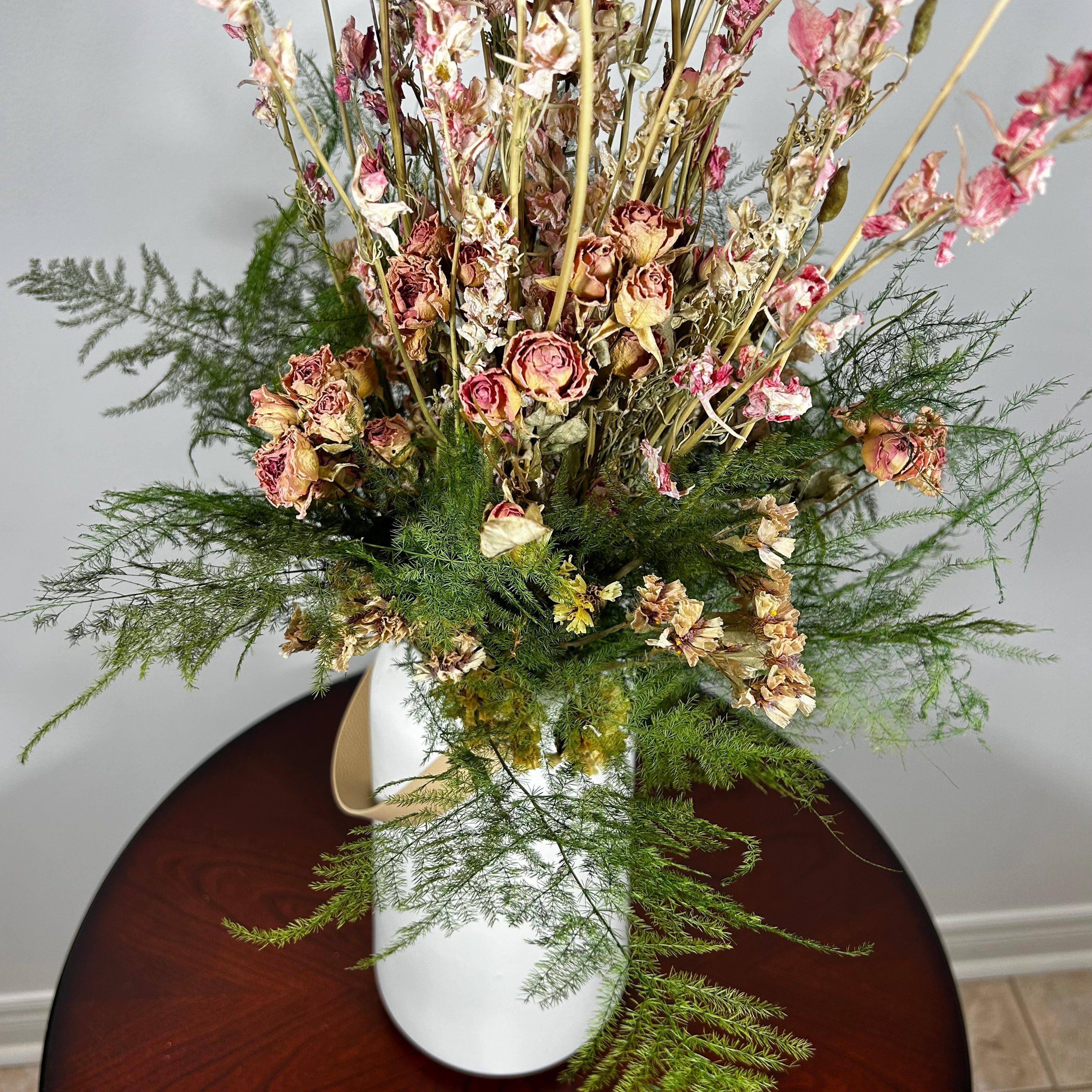 A white ceramic vase with a leather handle acts a the vessel from which a beautiful wisp of blooms rises. The central feature is the delicate spires of natural pink Larkspur, reaching upwards with gentle movement. These are complemented by charming clusters of natural pink roses and a base of lush, preserved asparagus greens. Cheerful accents of sunny yellow Statice complete this delightful composition, capturing the essence of a blossoming garden.