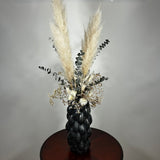 This dried floral arrangement has soft, elegant plumes of natural Pampas Grass reach gracefully upward, complemented by the bold, preserved forms of blue Eucalyptus and the airy lightness of German Statice (sea lavender). The delicate whimsy of bleached Triticum (wheat) wisps adds a touch of rustic charm, while soft white Cape Flowers provide subtle pops of pure, natural colour.