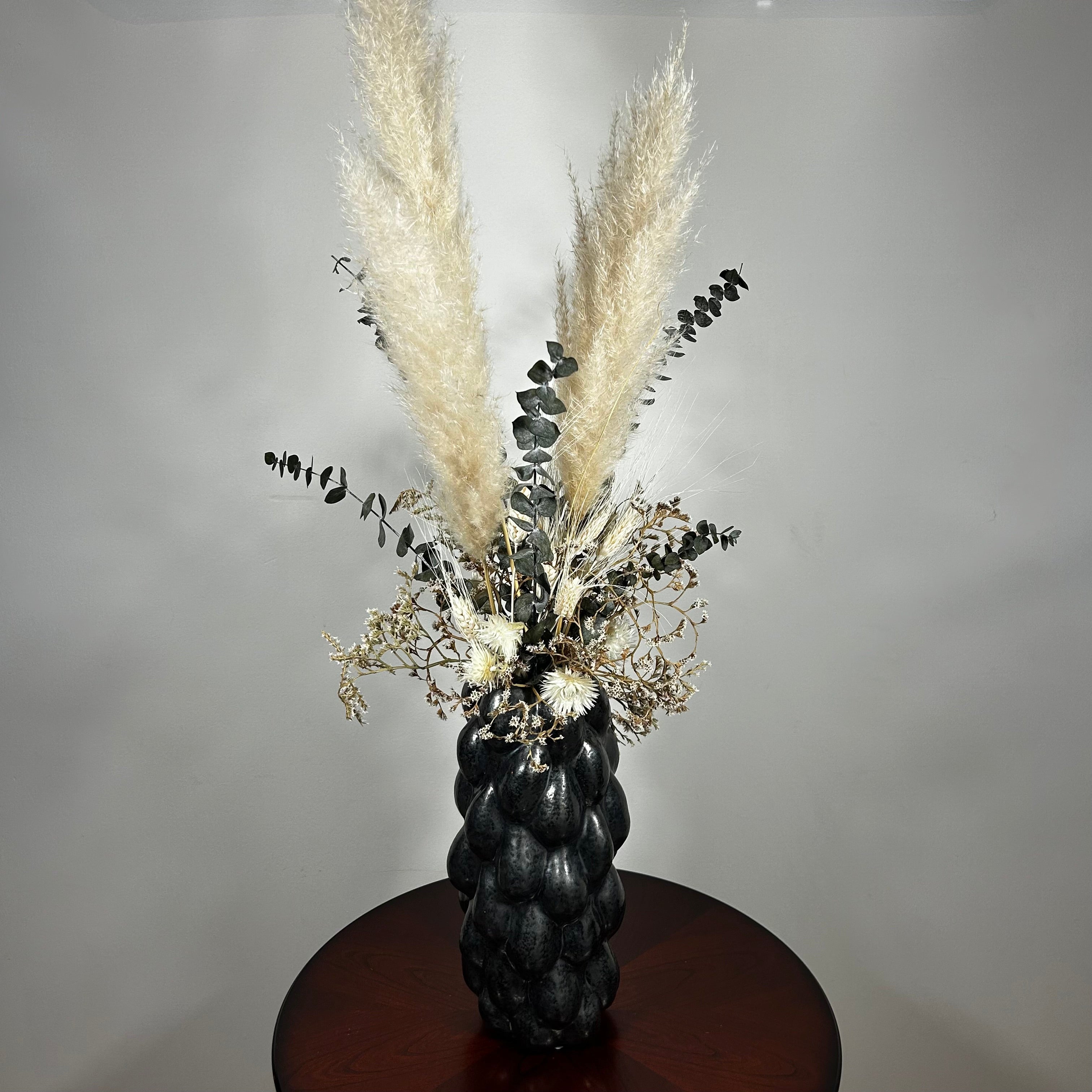 This dried floral arrangement has soft, elegant plumes of natural Pampas Grass reach gracefully upward, complemented by the bold, preserved forms of blue Eucalyptus and the airy lightness of German Statice (sea lavender). The delicate whimsy of bleached Triticum (wheat) wisps adds a touch of rustic charm, while soft white Cape Flowers provide subtle pops of pure, natural colour.