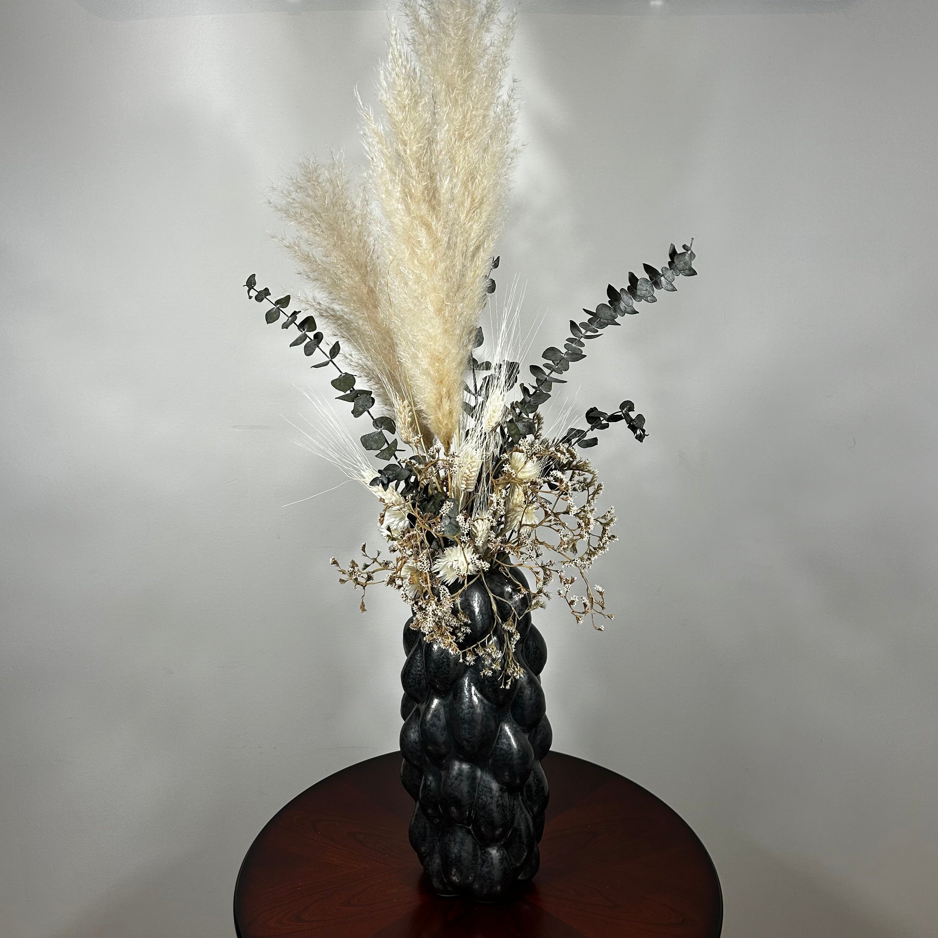This dried floral arrangement has soft, elegant plumes of natural Pampas Grass reach gracefully upward, complemented by the bold, preserved forms of blue Eucalyptus and the airy lightness of German Statice (sea lavender). The delicate whimsy of bleached Triticum (wheat) wisps adds a touch of rustic charm, while soft white Cape Flowers provide subtle pops of pure, natural colour.