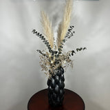 This dried floral arrangement has soft, elegant plumes of natural Pampas Grass reach gracefully upward, complemented by the bold, preserved forms of blue Eucalyptus and the airy lightness of German Statice (sea lavender). The delicate whimsy of bleached Triticum (wheat) wisps adds a touch of rustic charm, while soft white Cape Flowers provide subtle pops of pure, natural colour.