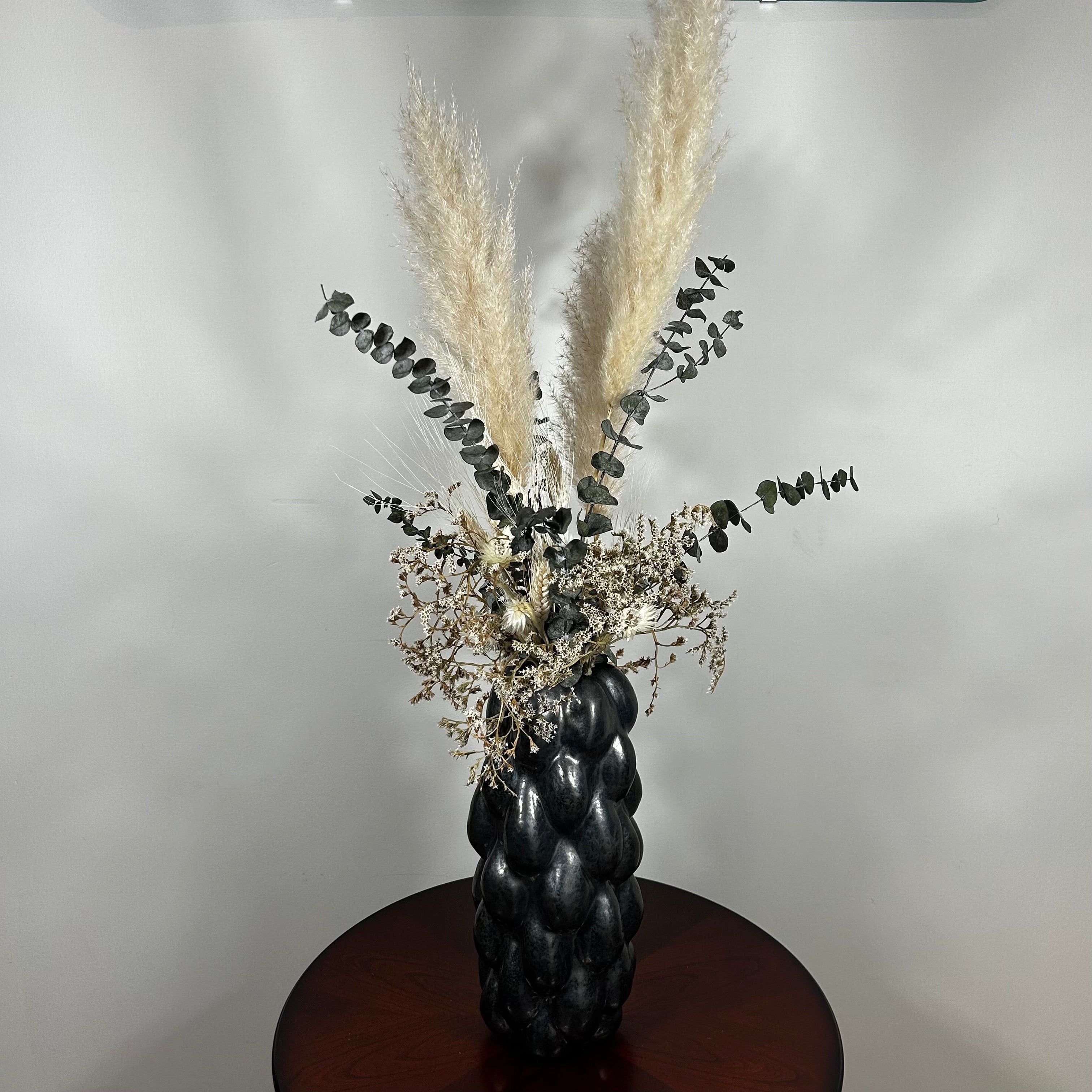 This dried floral arrangement has soft, elegant plumes of natural Pampas Grass reach gracefully upward, complemented by the bold, preserved forms of blue Eucalyptus and the airy lightness of German Statice (sea lavender). The delicate whimsy of bleached Triticum (wheat) wisps adds a touch of rustic charm, while soft white Cape Flowers provide subtle pops of pure, natural colour.