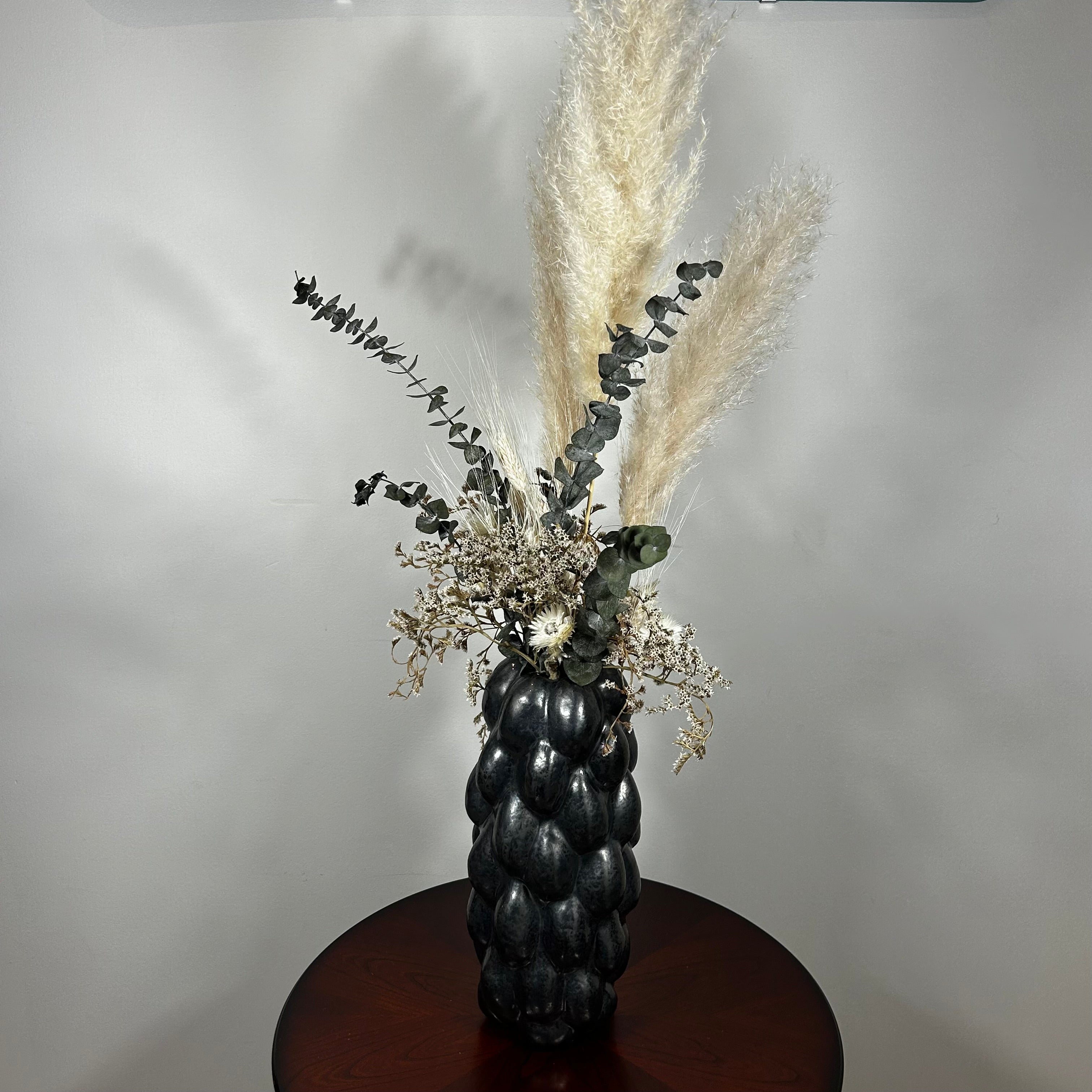 This dried floral arrangement has soft, elegant plumes of natural Pampas Grass reach gracefully upward, complemented by the bold, preserved forms of blue Eucalyptus and the airy lightness of German Statice (sea lavender). The delicate whimsy of bleached Triticum (wheat) wisps adds a touch of rustic charm, while soft white Cape Flowers provide subtle pops of pure, natural colour.