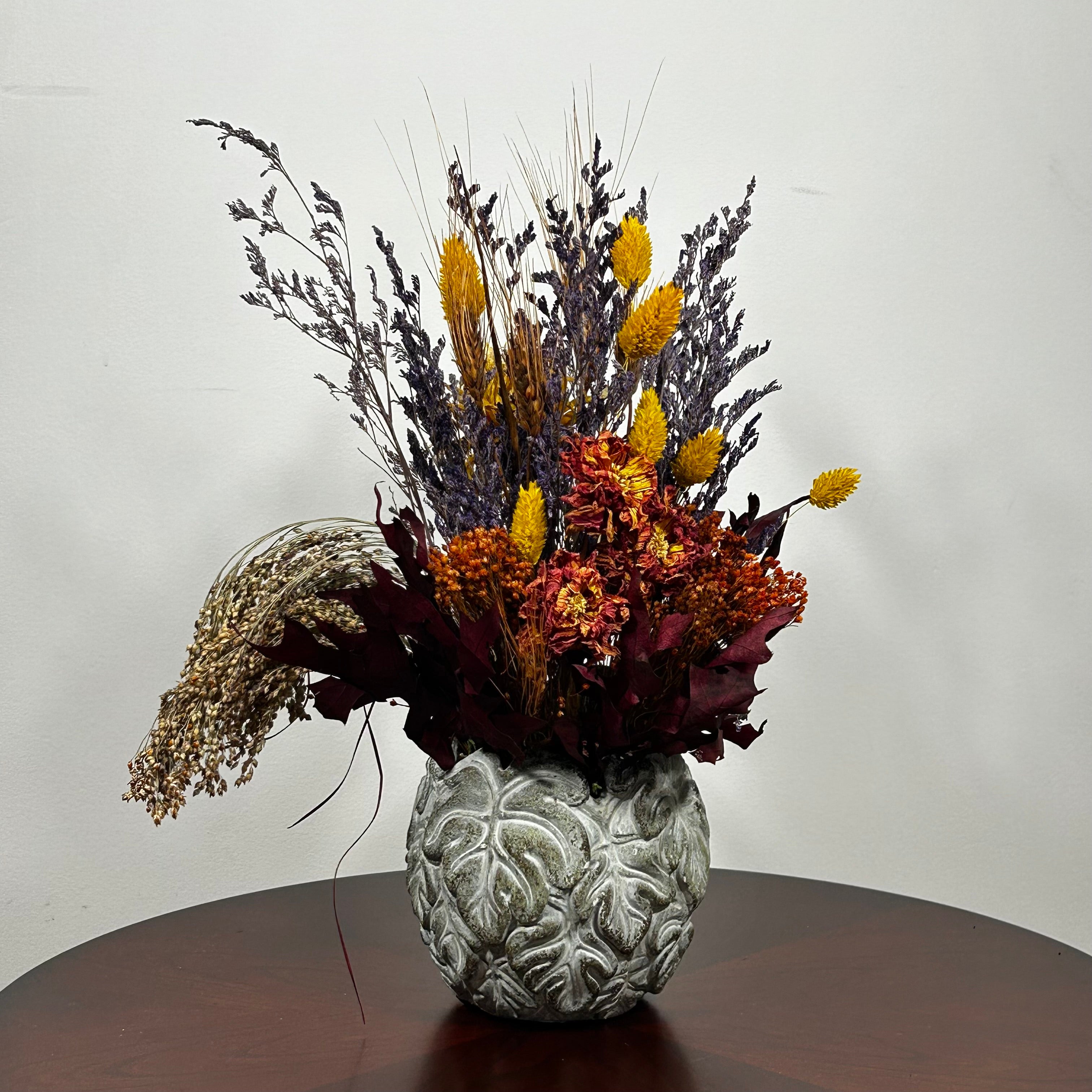 Showcasing a dried floral arrangement in a concrete vessel shaped like a wreath made out of jungle leaves. Filled  with warm orange Achillea Parker (Yarrow) reaches upwards, complemented by the delicate spires of super blue Lavender. A sense of abundance is added with dried Sorghum (Milo) in a deep orange and slender dried Wheat Stalks in a rich chocolate brown. The entire composition is finished with playful bursts of golden yellow Phalaris (Canary Grass), ensuring a luminous, sun-kissed feel.