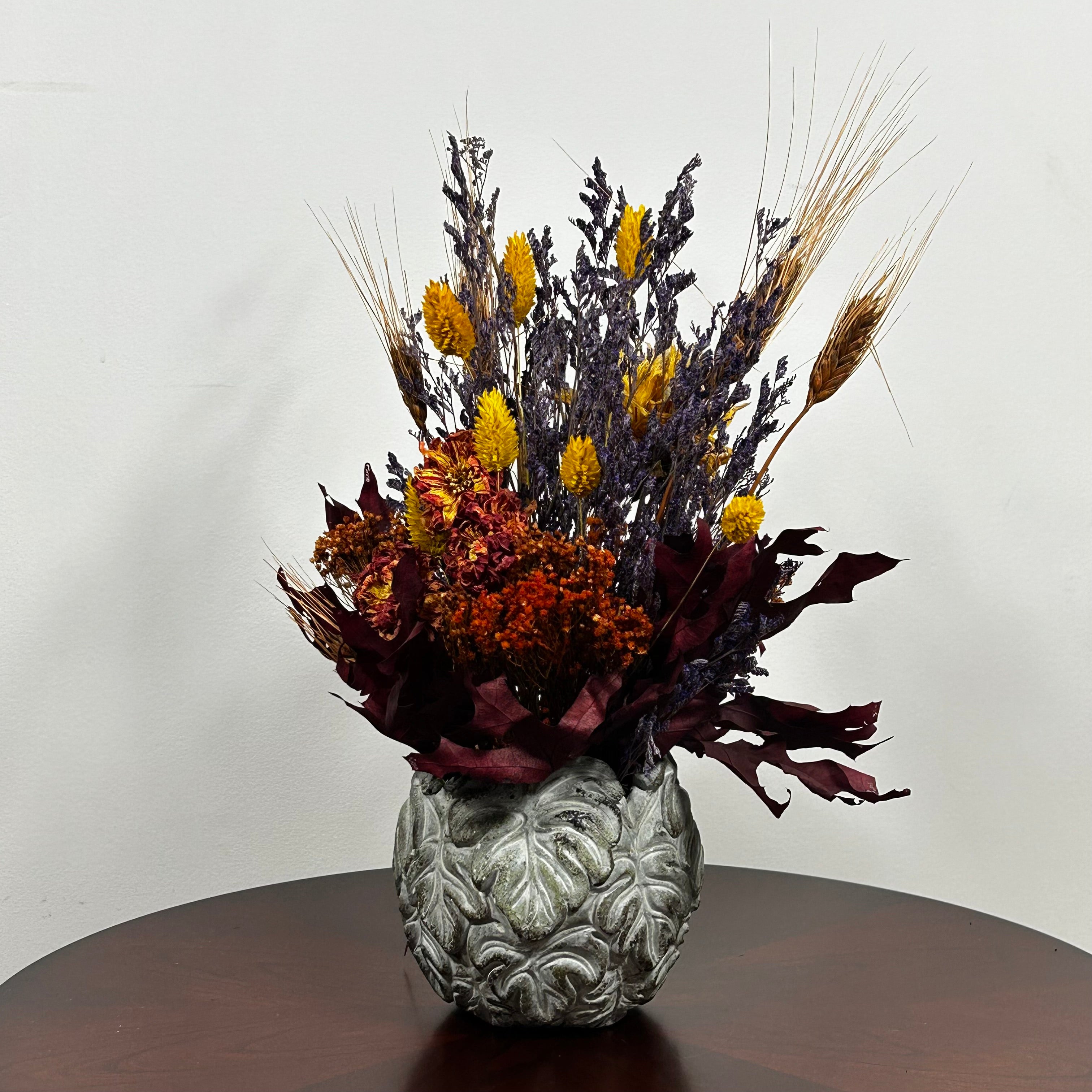 Showcasing a dried floral arrangement in a concrete vessel shaped like a wreath made out of jungle leaves. Filled  with warm orange Achillea Parker (Yarrow) reaches upwards, complemented by the delicate spires of super blue Lavender. A sense of abundance is added with dried Sorghum (Milo) in a deep orange and slender dried Wheat Stalks in a rich chocolate brown. The entire composition is finished with playful bursts of golden yellow Phalaris (Canary Grass), ensuring a luminous, sun-kissed feel.
