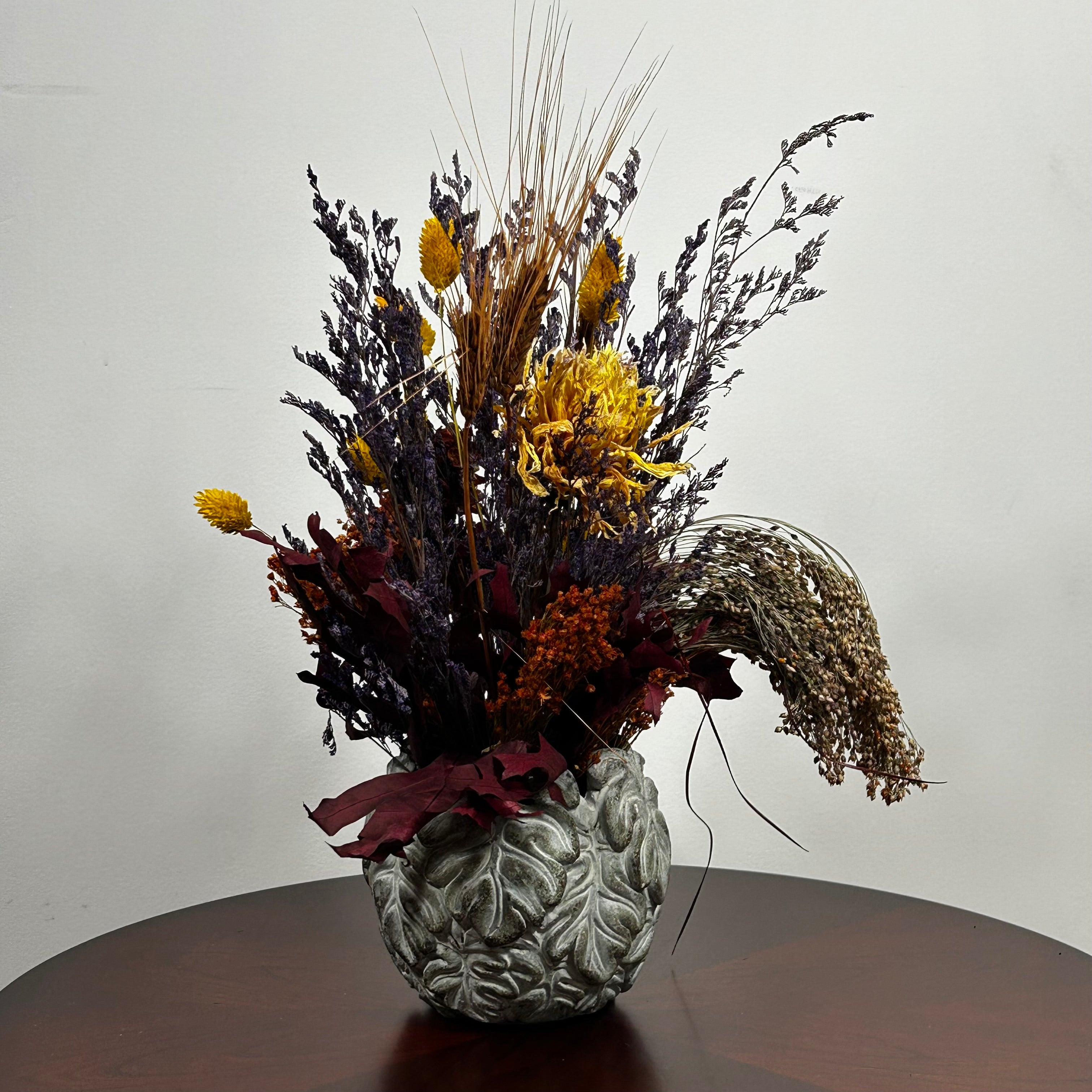 Showcasing a dried floral arrangement in a concrete vessel shaped like a wreath made out of jungle leaves. Filled  with warm orange Achillea Parker (Yarrow) reaches upwards, complemented by the delicate spires of super blue Lavender. A sense of abundance is added with dried Sorghum (Milo) in a deep orange and slender dried Wheat Stalks in a rich chocolate brown. The entire composition is finished with playful bursts of golden yellow Phalaris (Canary Grass), ensuring a luminous, sun-kissed feel.