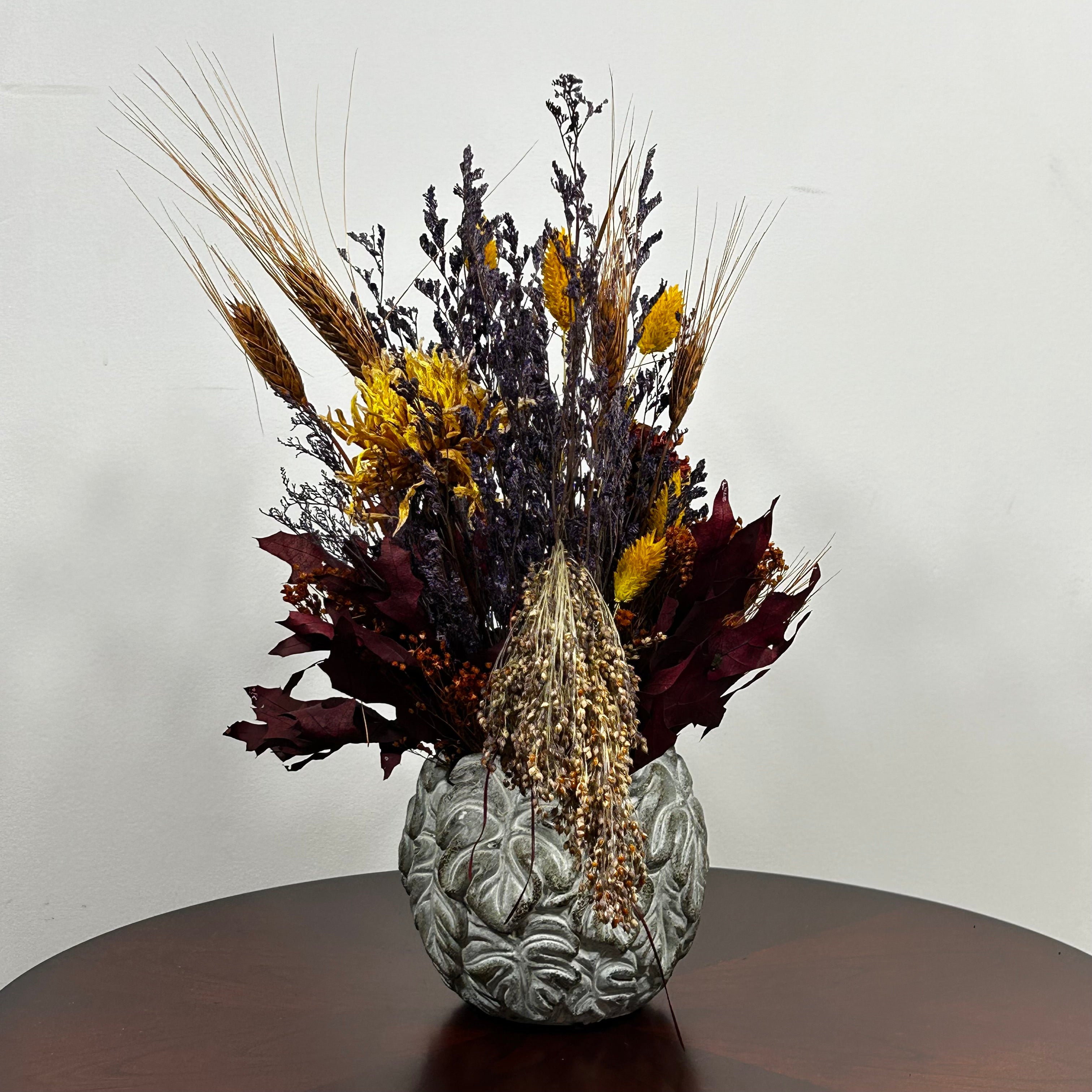 Showcasing a dried floral arrangement in a concrete vessel shaped like a wreath made out of jungle leaves. Filled  with warm orange Achillea Parker (Yarrow) reaches upwards, complemented by the delicate spires of super blue Lavender. A sense of abundance is added with dried Sorghum (Milo) in a deep orange and slender dried Wheat Stalks in a rich chocolate brown. The entire composition is finished with playful bursts of golden yellow Phalaris (Canary Grass), ensuring a luminous, sun-kissed feel.