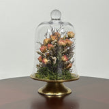 The floral composition features lovely soft pink spray roses, blending harmoniously with the subtle textures of German Statice (sea lavender) and the delicate purple of caspia lavender. Spanish moss provides a soft, textural foundation, completing this miniature world of preserved loveliness. It is built on an antiqued gold pedestal covered by a clear glass cloche. 