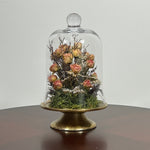 The floral composition features lovely soft pink spray roses, blending harmoniously with the subtle textures of German Statice (sea lavender) and the delicate purple of caspia lavender. Spanish moss provides a soft, textural foundation, completing this miniature world of preserved loveliness. It is built on an antiqued gold pedestal covered by a clear glass cloche. 