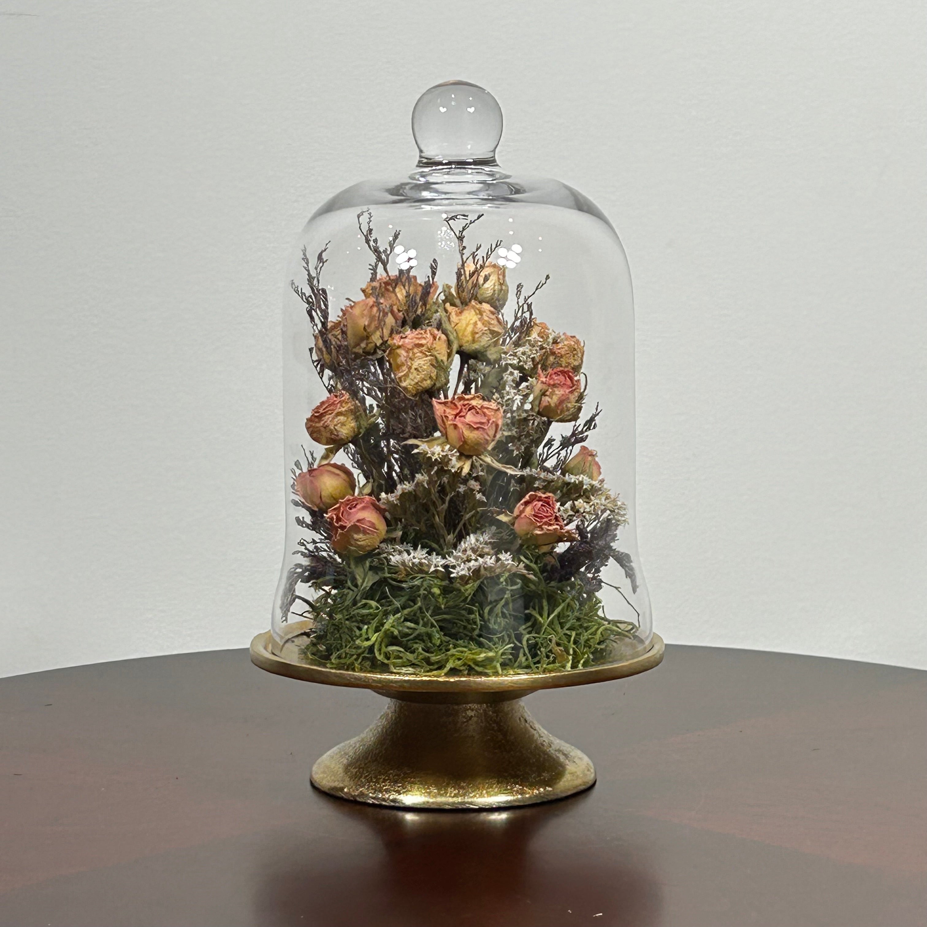 The floral composition features lovely soft pink spray roses, blending harmoniously with the subtle textures of German Statice (sea lavender) and the delicate purple of caspia lavender. Spanish moss provides a soft, textural foundation, completing this miniature world of preserved loveliness. It is built on an antiqued gold pedestal covered by a clear glass cloche. 