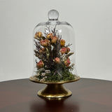 The floral composition features lovely soft pink spray roses, blending harmoniously with the subtle textures of German Statice (sea lavender) and the delicate purple of caspia lavender. Spanish moss provides a soft, textural foundation, completing this miniature world of preserved loveliness. It is built on an antiqued gold pedestal covered by a clear glass cloche. 