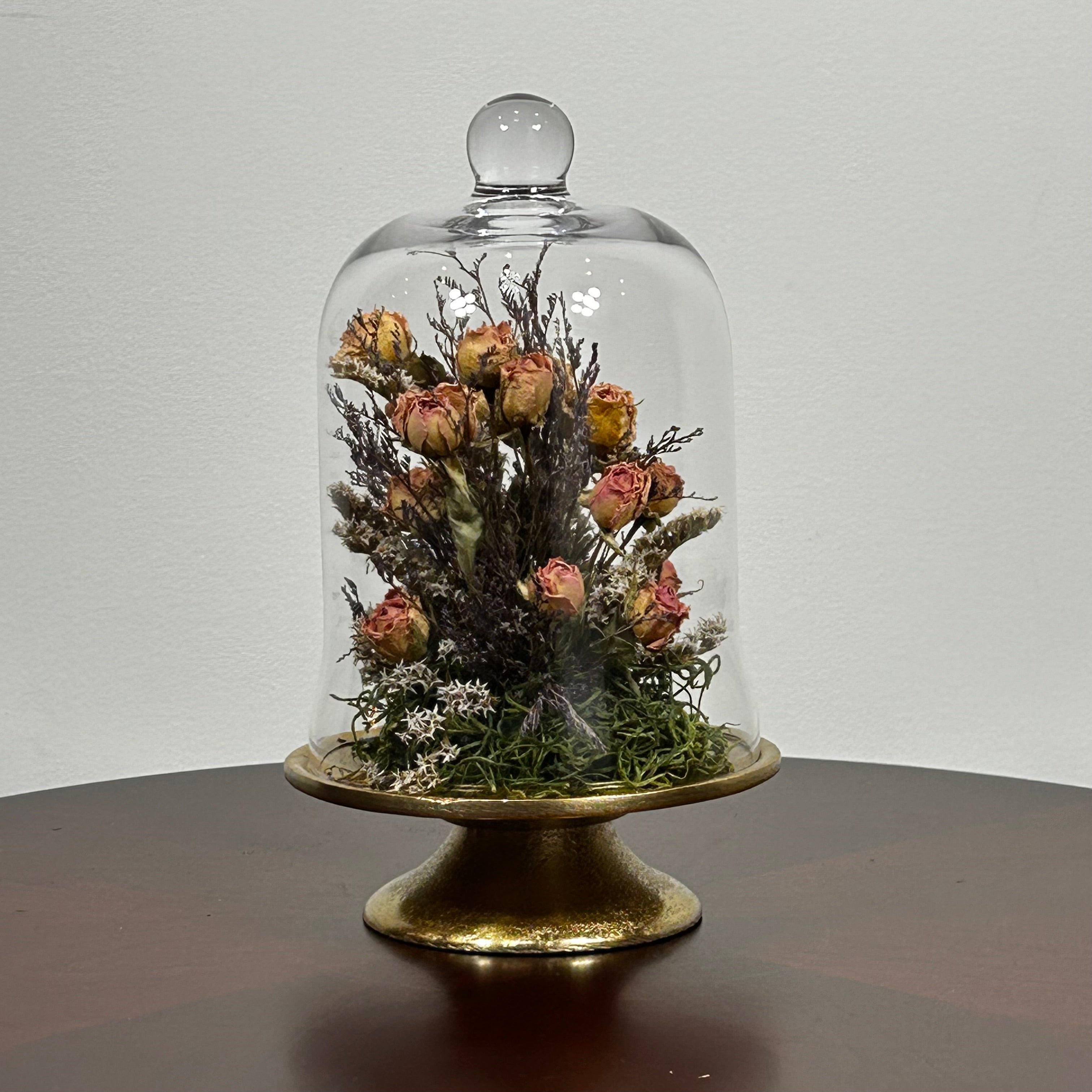The floral composition features lovely soft pink spray roses, blending harmoniously with the subtle textures of German Statice (sea lavender) and the delicate purple of caspia lavender. Spanish moss provides a soft, textural foundation, completing this miniature world of preserved loveliness. It is built on an antiqued gold pedestal covered by a clear glass cloche. 