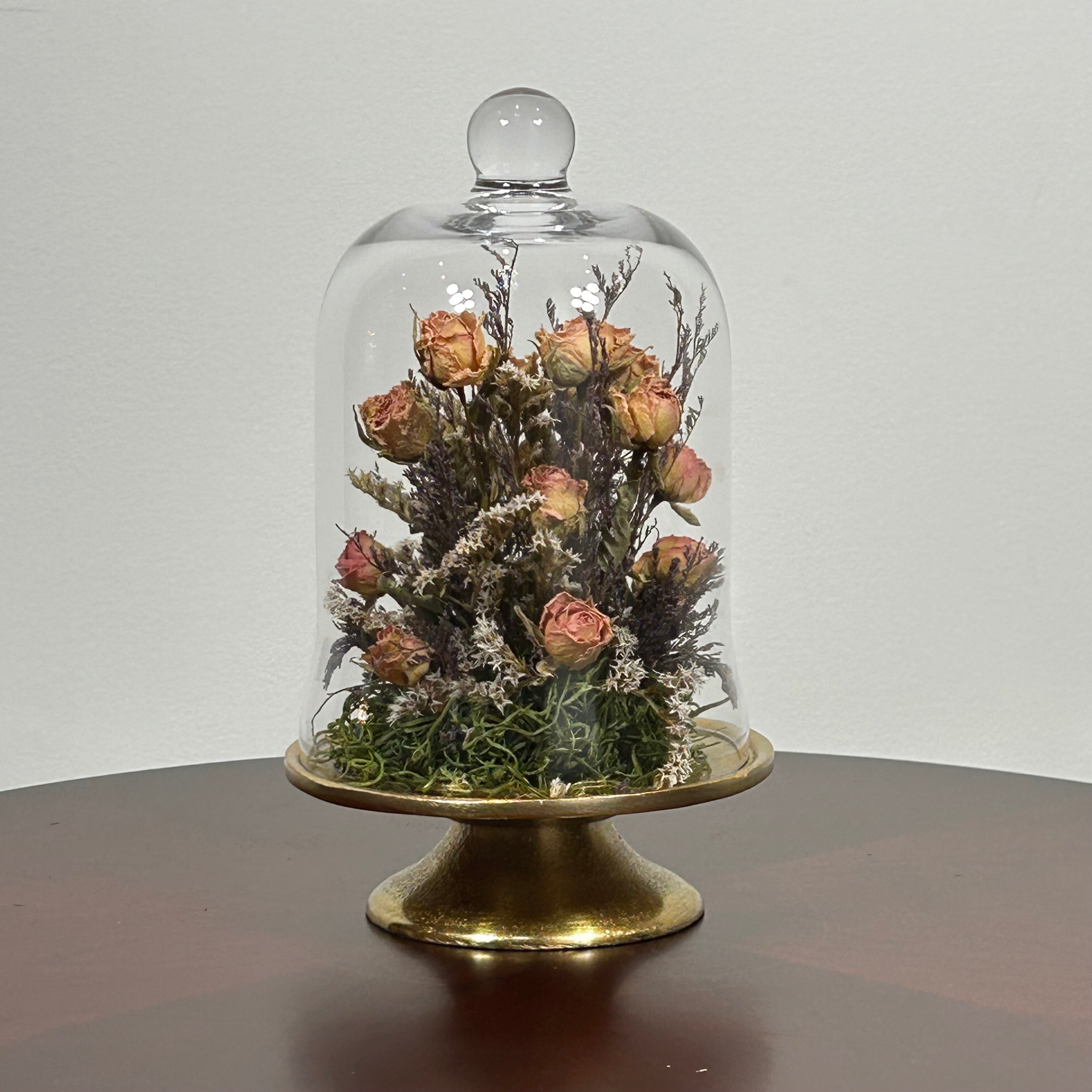 The floral composition features lovely soft pink spray roses, blending harmoniously with the subtle textures of German Statice (sea lavender) and the delicate purple of caspia lavender. Spanish moss provides a soft, textural foundation, completing this miniature world of preserved loveliness. It is built on an antiqued gold pedestal covered by a clear glass cloche. 