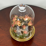 The floral composition features lovely soft pink spray roses, blending harmoniously with the subtle textures of German Statice (sea lavender) and the delicate purple of caspia lavender. Spanish moss provides a soft, textural foundation, completing this miniature world of preserved loveliness. It is built on an antiqued gold pedestal covered by a clear glass cloche. 