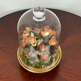 The floral composition features lovely soft pink spray roses, blending harmoniously with the subtle textures of German Statice (sea lavender) and the delicate purple of caspia lavender. Spanish moss provides a soft, textural foundation, completing this miniature world of preserved loveliness. It is built on an antiqued gold pedestal covered by a clear glass cloche. 