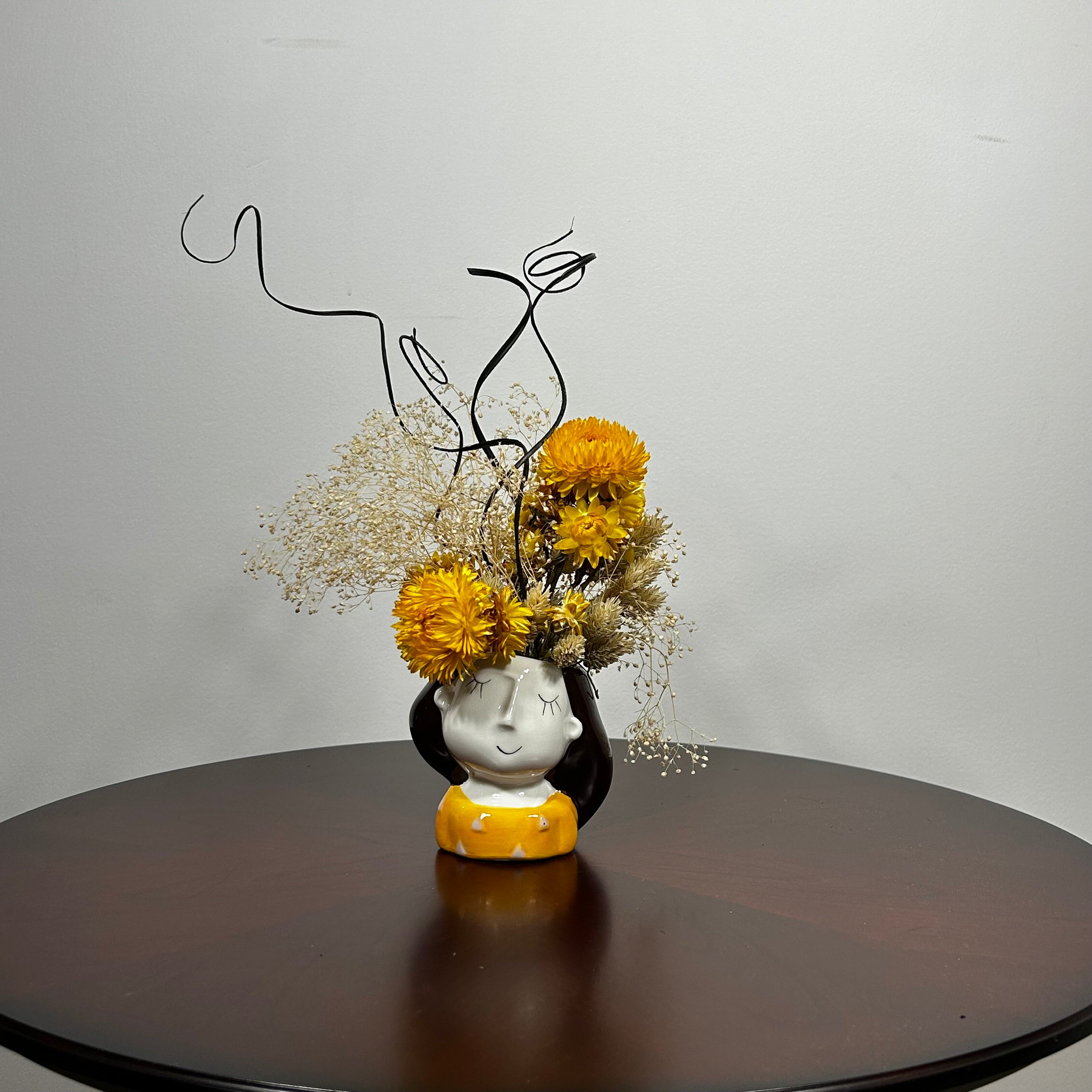 A unique ceramic vessel, sculpted as a demure woman's head adorned with rich brown hair and a striking golden blouse hosts a radiant array of everlasting blooms. The design features luminous golden yellow Helichrysum (strawflowers), delicate Carolina canary grass, and airy Gypsophila(baby's breath). Playful curly ting ting is added to lend a final touch of whimsy and texture to her crown. 