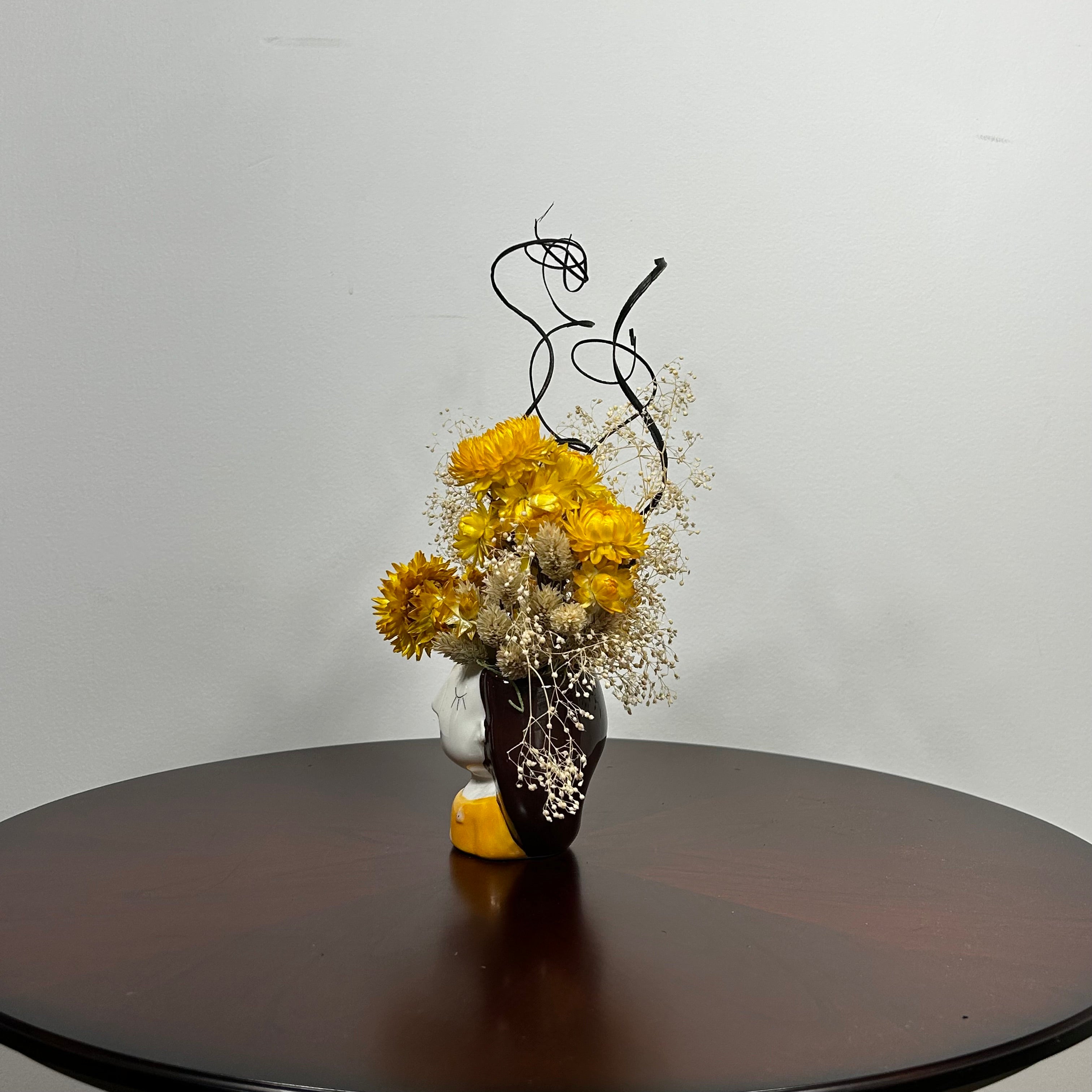 A unique ceramic vessel, sculpted as a demure woman's head adorned with rich brown hair and a striking golden blouse hosts a radiant array of everlasting blooms. The design features luminous golden yellow Helichrysum (strawflowers), delicate Carolina canary grass, and airy Gypsophila(baby's breath). Playful curly ting ting is added to lend a final touch of whimsy and texture to her crown. 