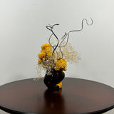 A unique ceramic vessel, sculpted as a demure woman's head adorned with rich brown hair and a striking golden blouse hosts a radiant array of everlasting blooms. The design features luminous golden yellow Helichrysum (strawflowers), delicate Carolina canary grass, and airy Gypsophila(baby's breath). Playful curly ting ting is added to lend a final touch of whimsy and texture to her crown. 
