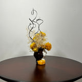 A unique ceramic vessel, sculpted as a demure woman's head adorned with rich brown hair and a striking golden blouse hosts a radiant array of everlasting blooms. The design features luminous golden yellow Helichrysum (strawflowers), delicate Carolina canary grass, and airy Gypsophila(baby's breath). Playful curly ting ting is added to lend a final touch of whimsy and texture to her crown. 