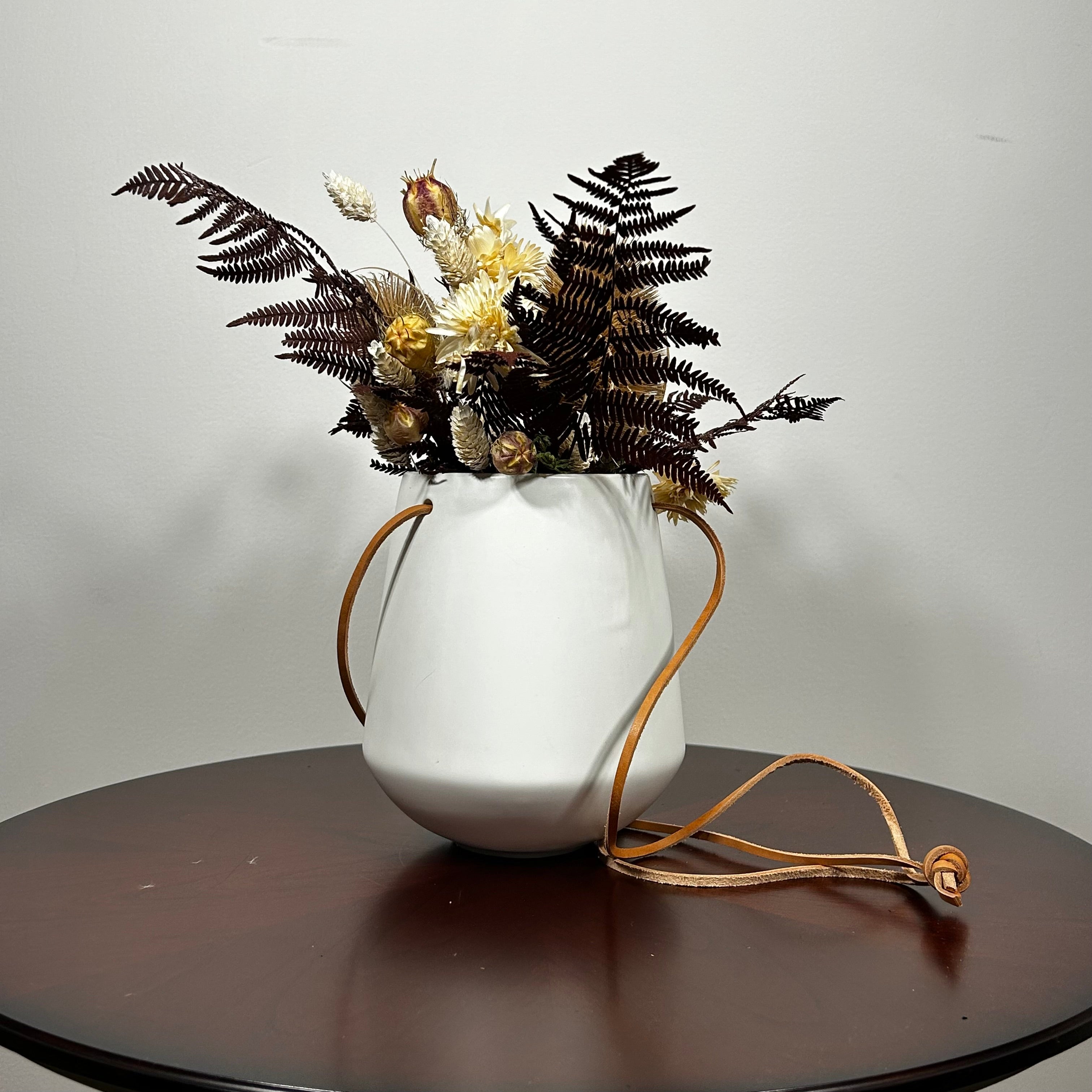 White hanging planter with beautiful dried flowers arrangement with burgundy bracken, ivory helichrysums, Nigel seed pods, natural teasels, and ivory phalaris on a beige background