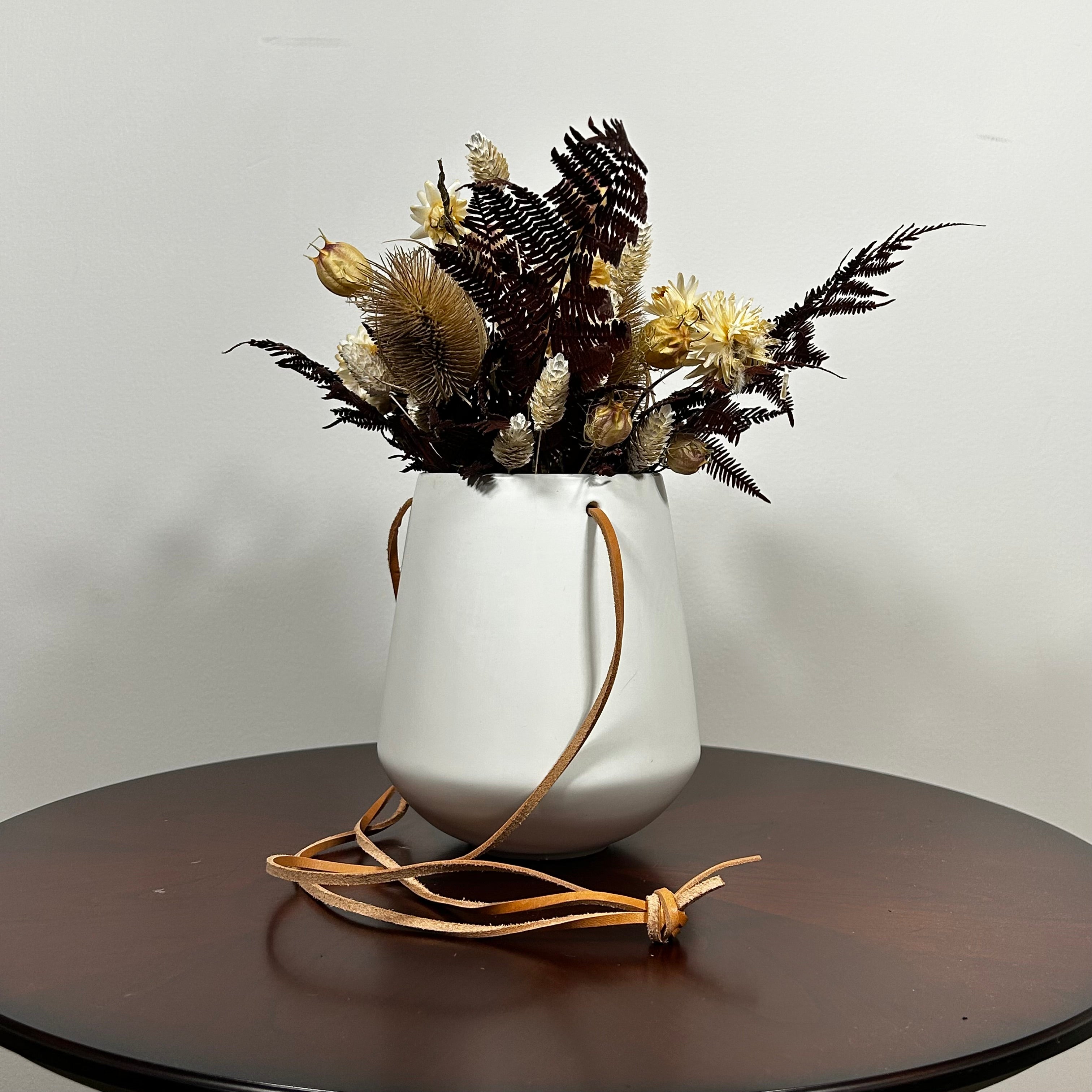 White hanging planter with beautiful dried flowers arrangement with burgundy bracken, ivory helichrysums, Nigel seed pods, natural teasels, and ivory phalaris on a beige background